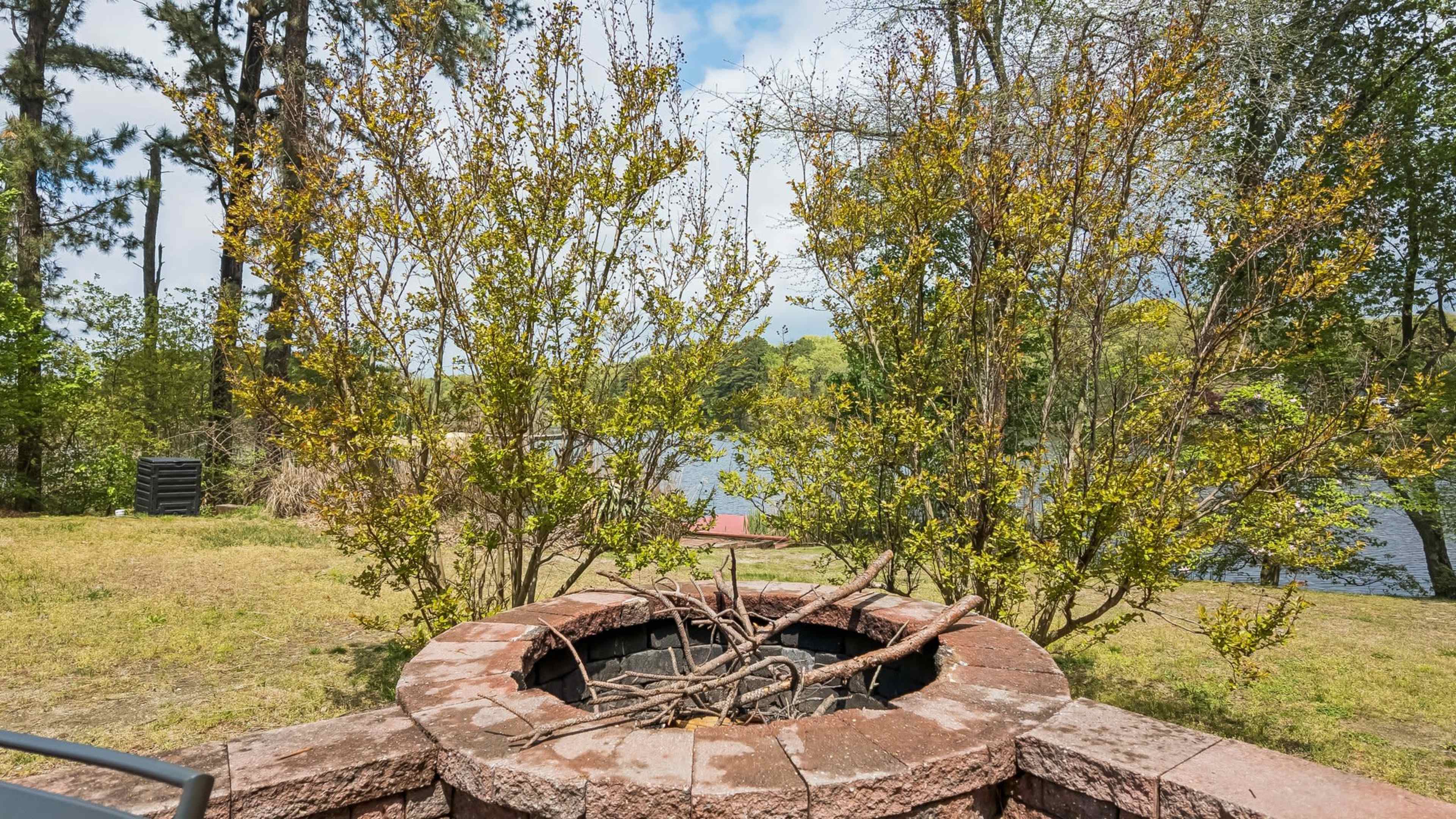 A fire pit surrounded by bushes and trees overlooks a tranquil body of water.