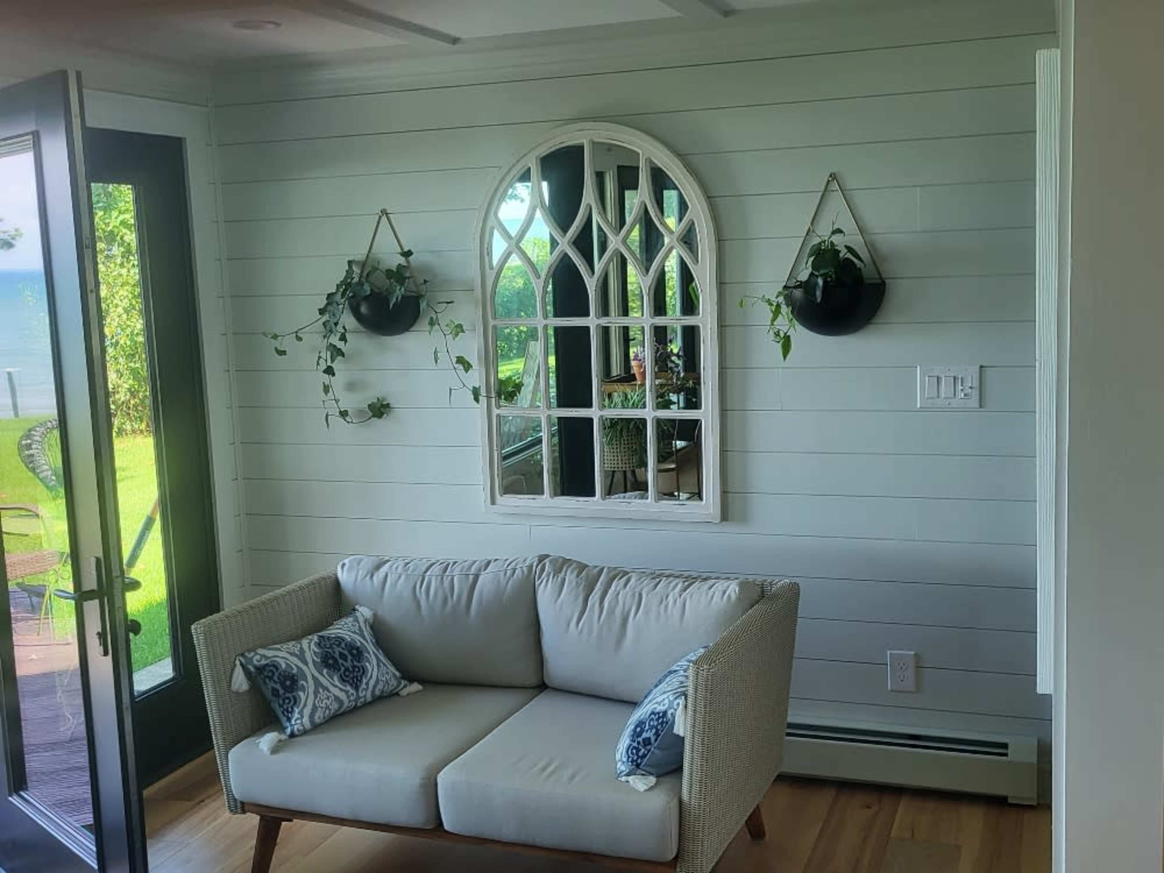 A light-colored living space featuring a sofa with decorative pillows, a wall mirror, and greenery in hanging planters, with large windows revealing an outdoor view.