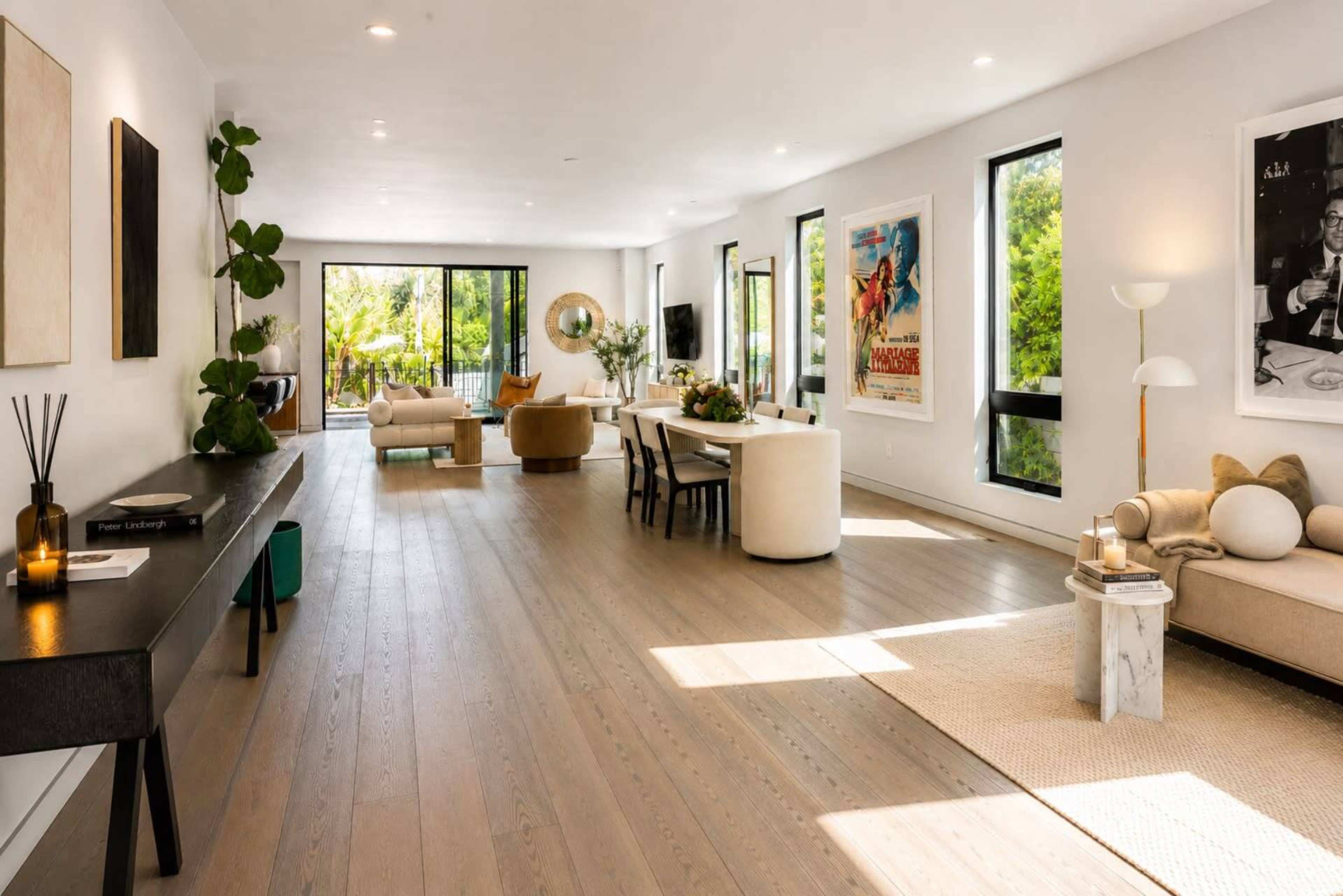 The image shows a spacious, modern living room with large windows, hardwood floors, and contemporary furniture arranged for socializing.