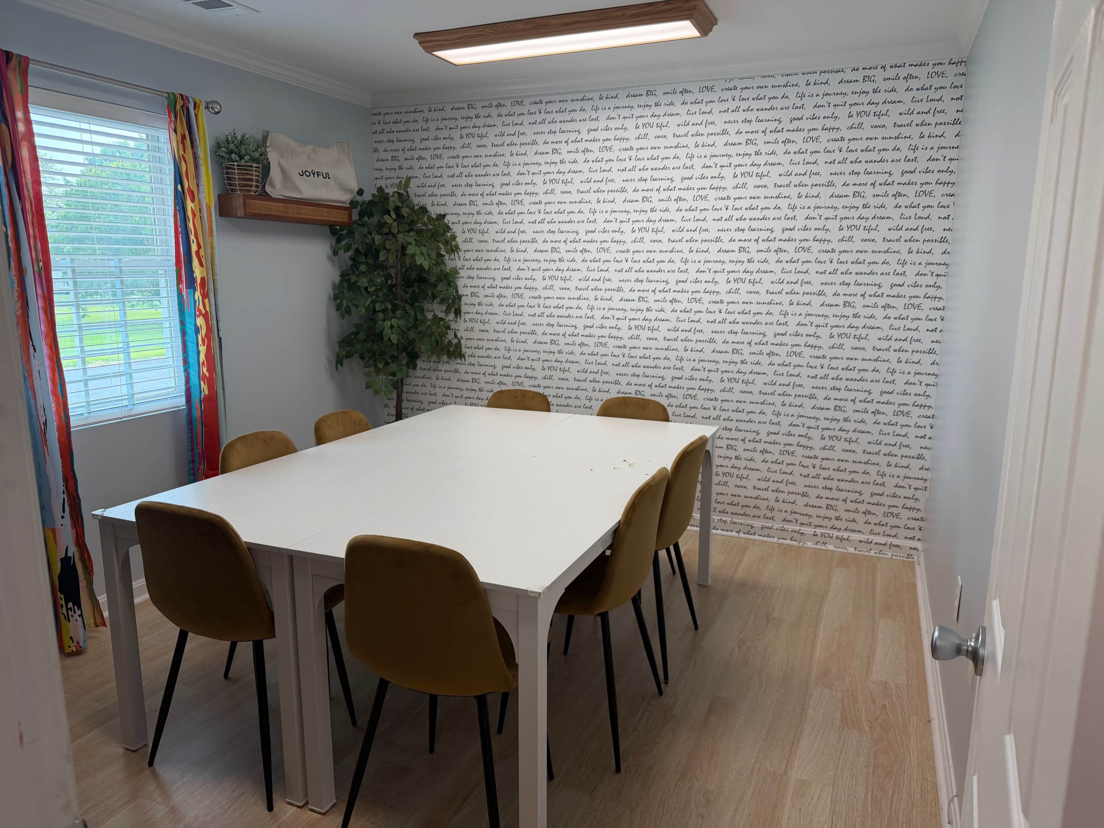 A modern meeting room features a large white table surrounded by six tan chairs, with a wall adorned with text and a small plant near a window.