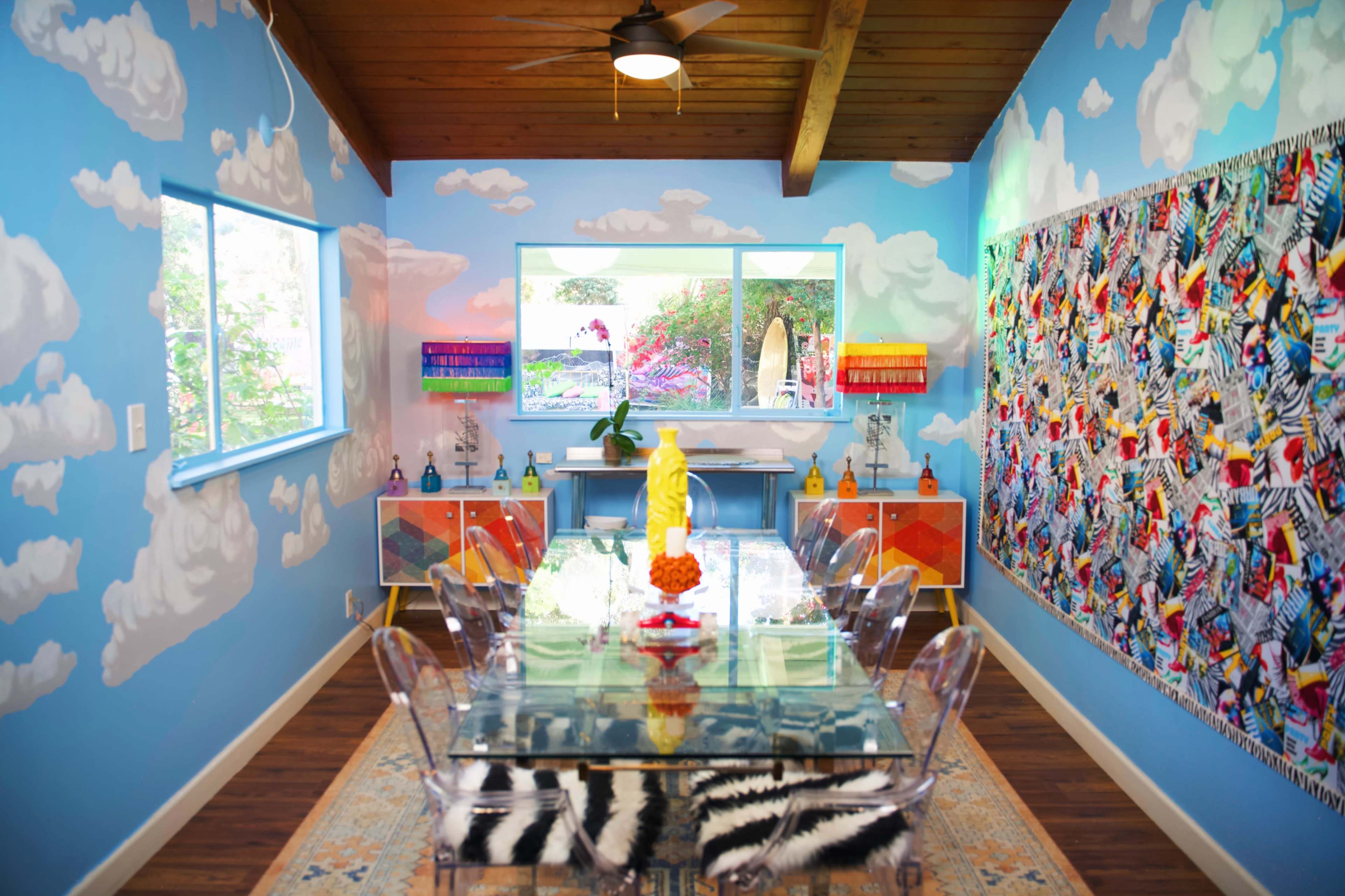 A brightly colored dining room features a clear table surrounded by transparent chairs, with cloud-painted walls and a vibrant, abstract art piece on one side.