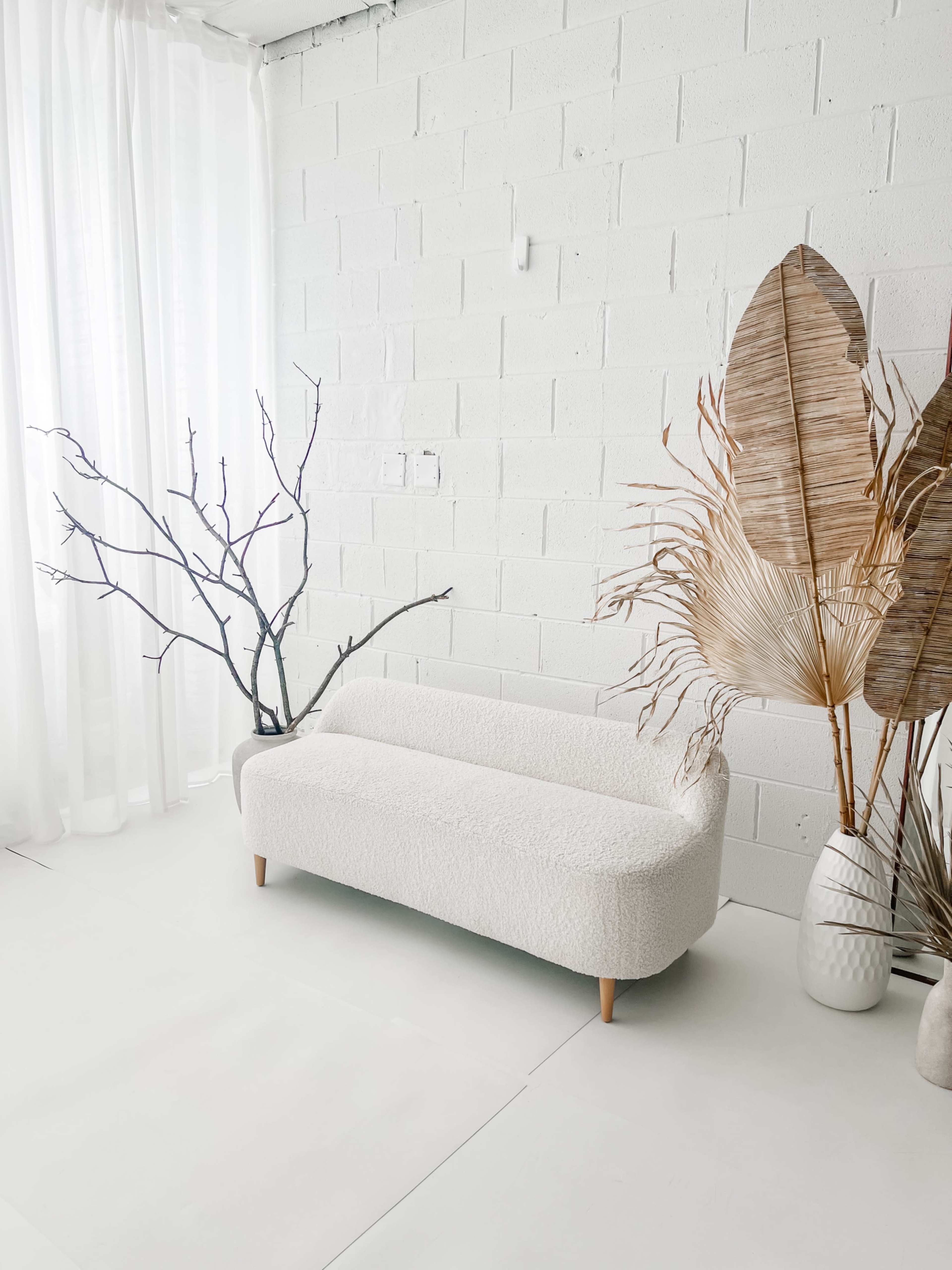 A white, textured couch is positioned in a bright room next to a tall, bare branch and large dried leaves in vases against a white wall.