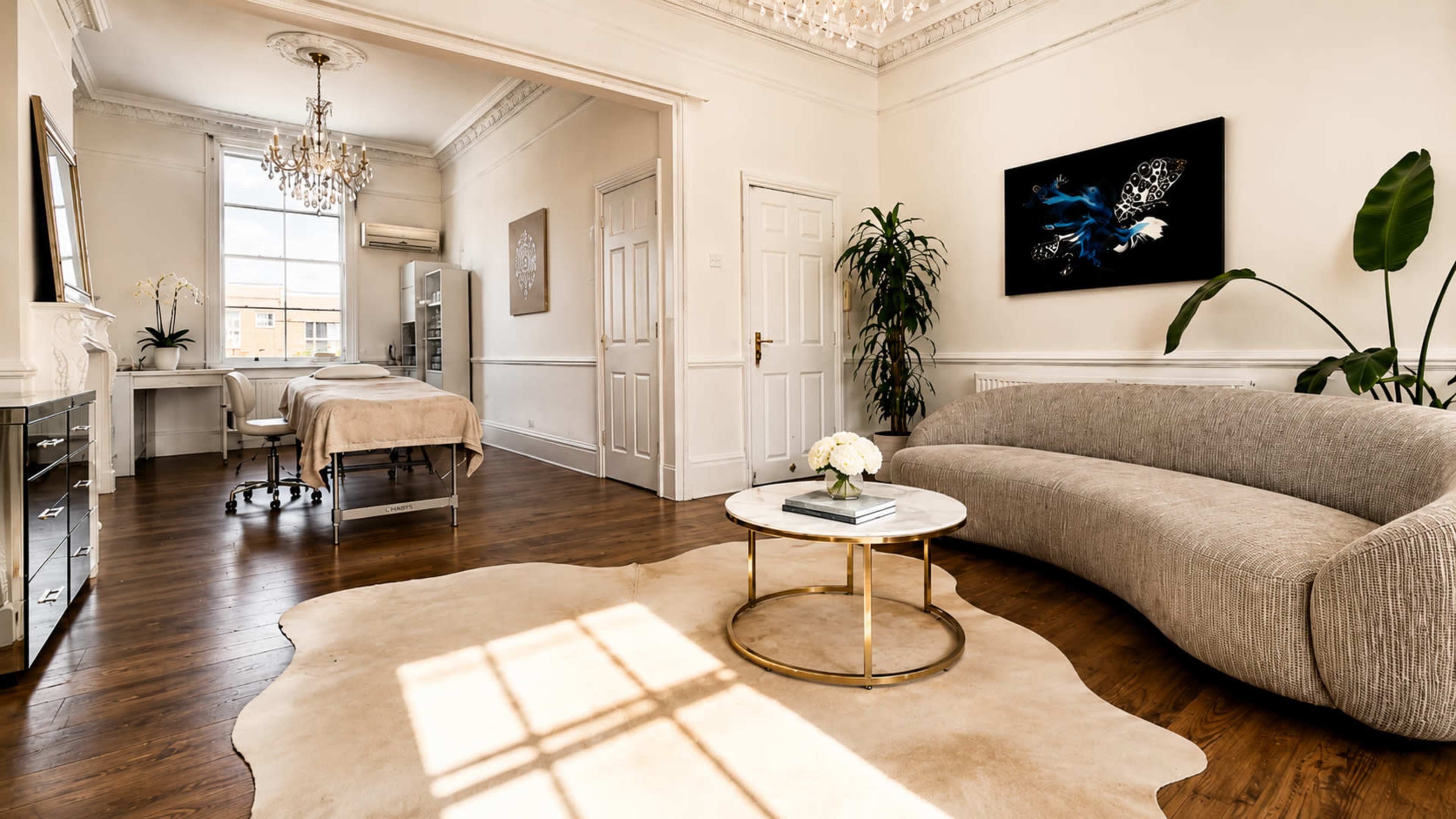 The image shows a well-lit treatment room featuring a cozy couch, a round coffee table, a treatment bed, and a large window casting shadows on the wooden floor.