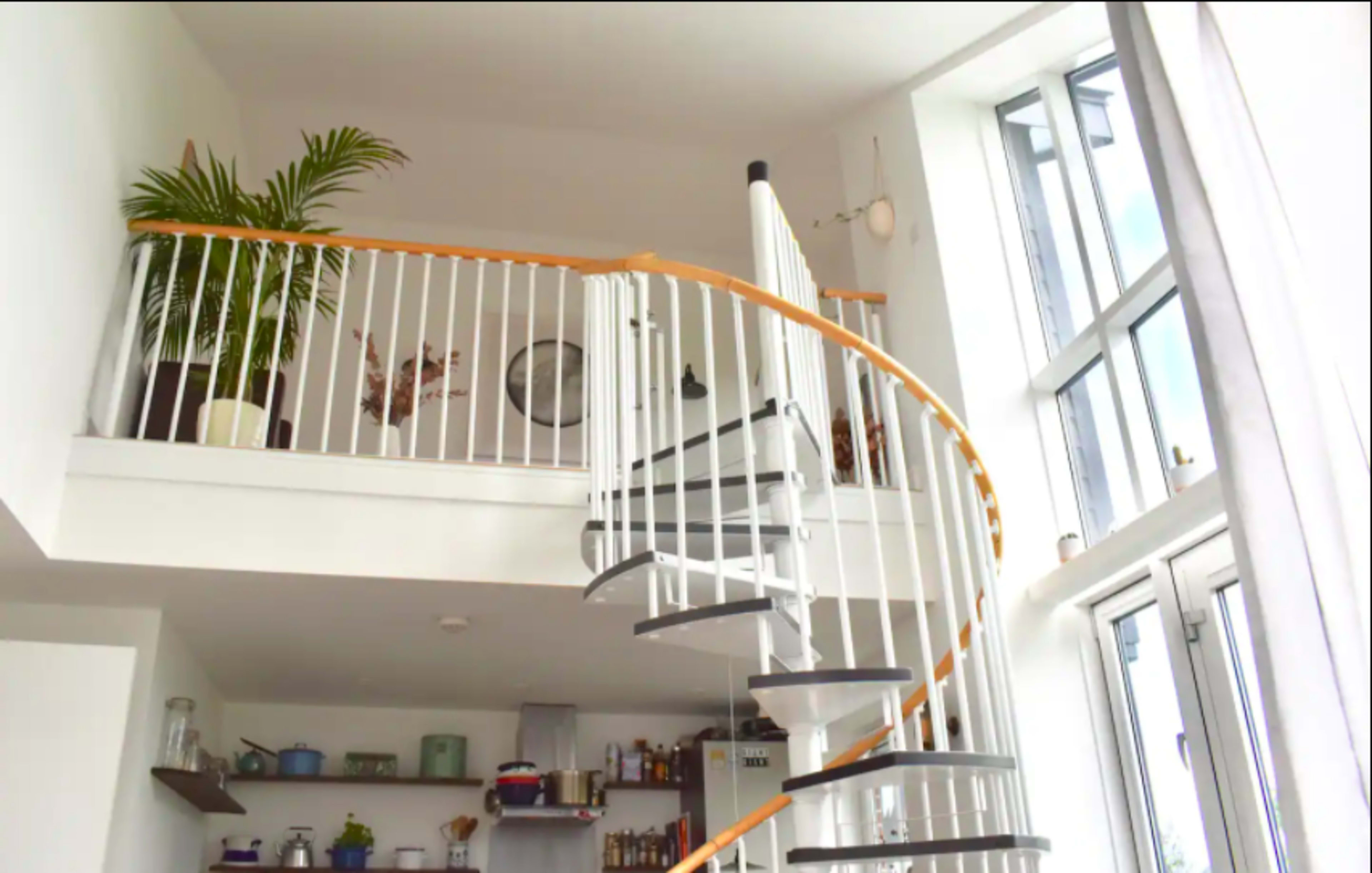 The image shows a modern interior featuring a white spiral staircase leading to a loft space, with large windows and greenery visible in the atrium.