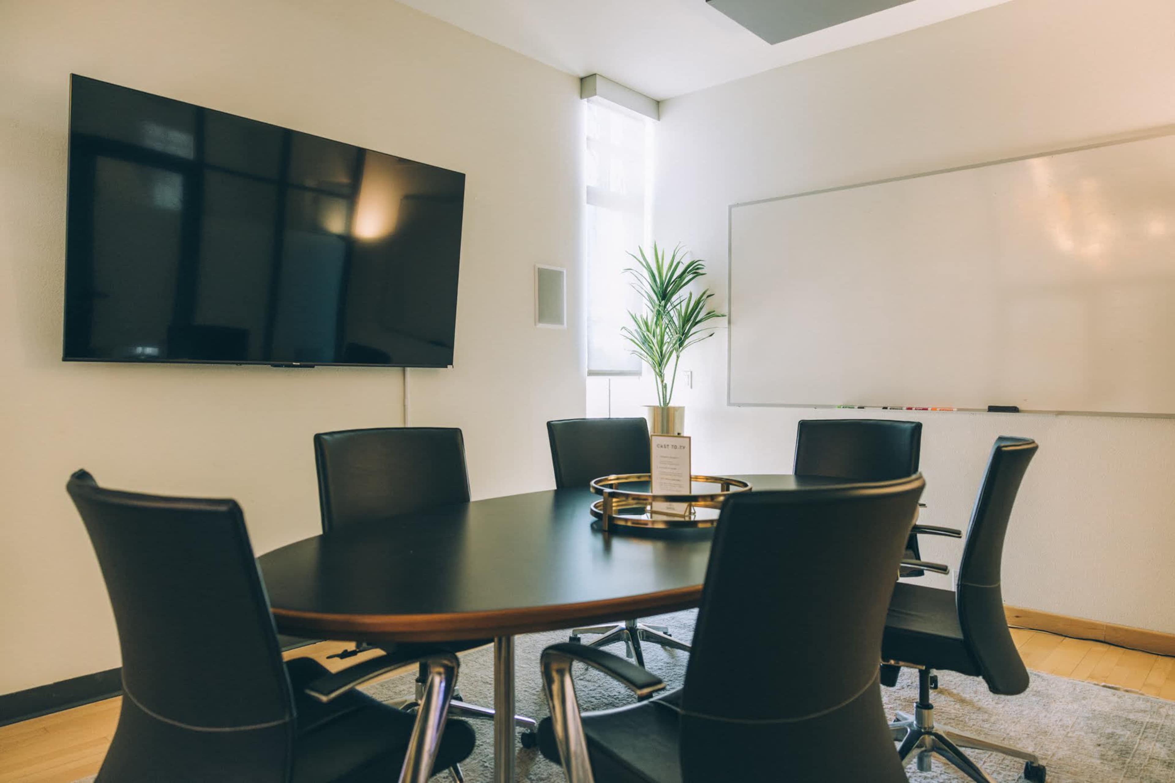 A modern conference room features a large television, a whiteboard, a round table, and six black chairs arranged around it, with a small decorative plant in the center.