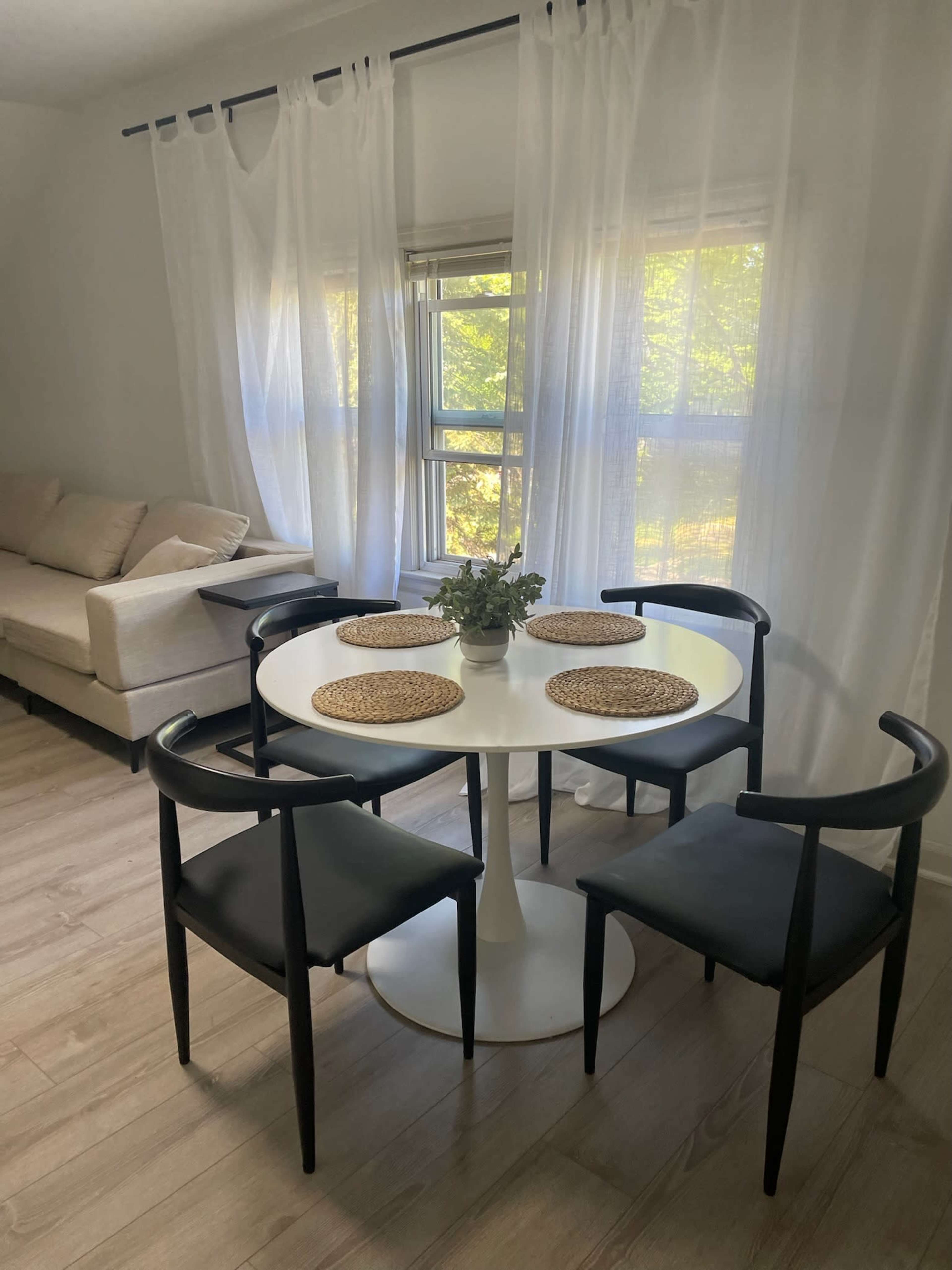 A round white dining table with black chairs is set with four placemats in a bright room featuring sheer white curtains and a beige sofa.