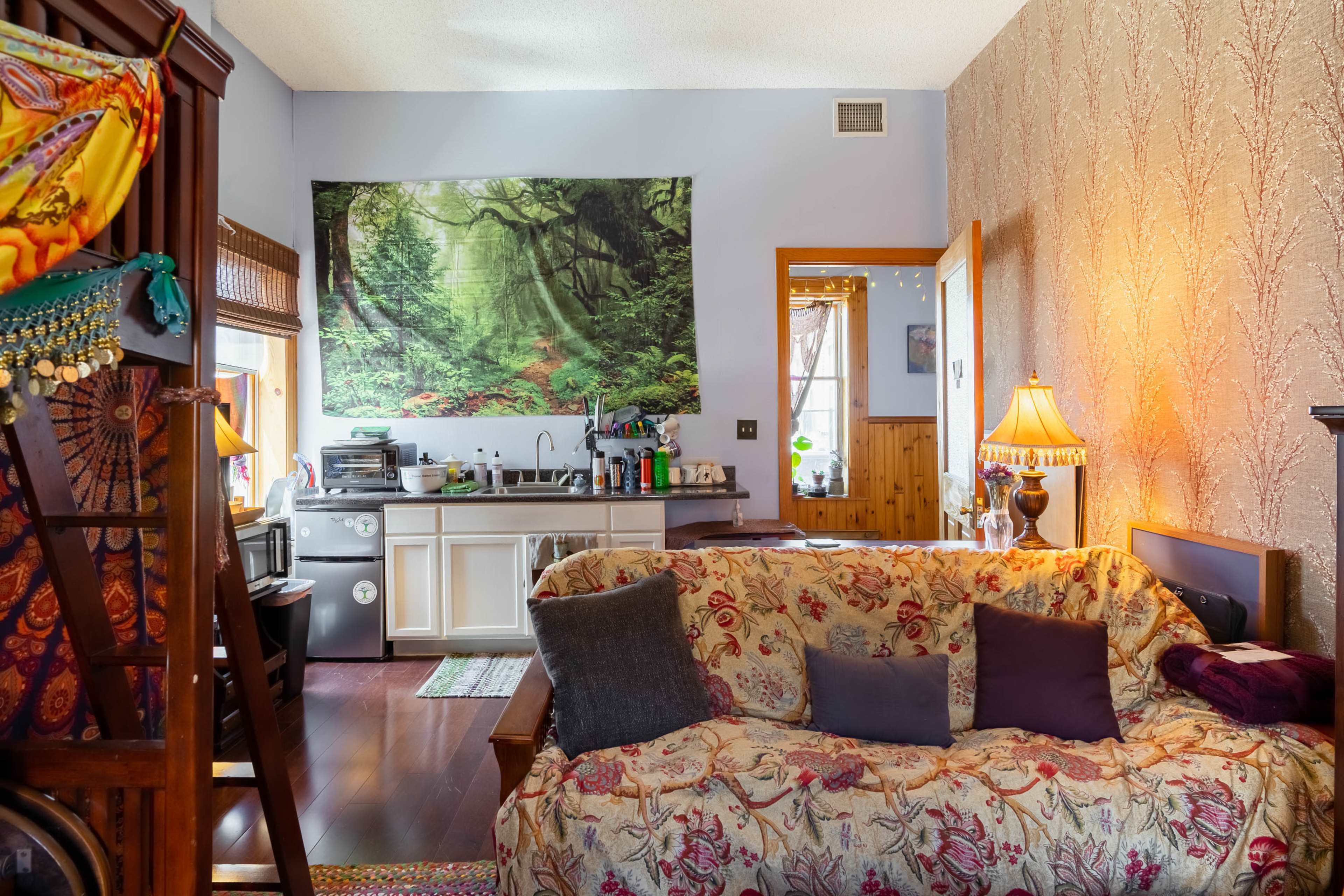 The image shows a small living area with a couch, a kitchenette in the background, and a large wall tapestry depicting a forest scene.