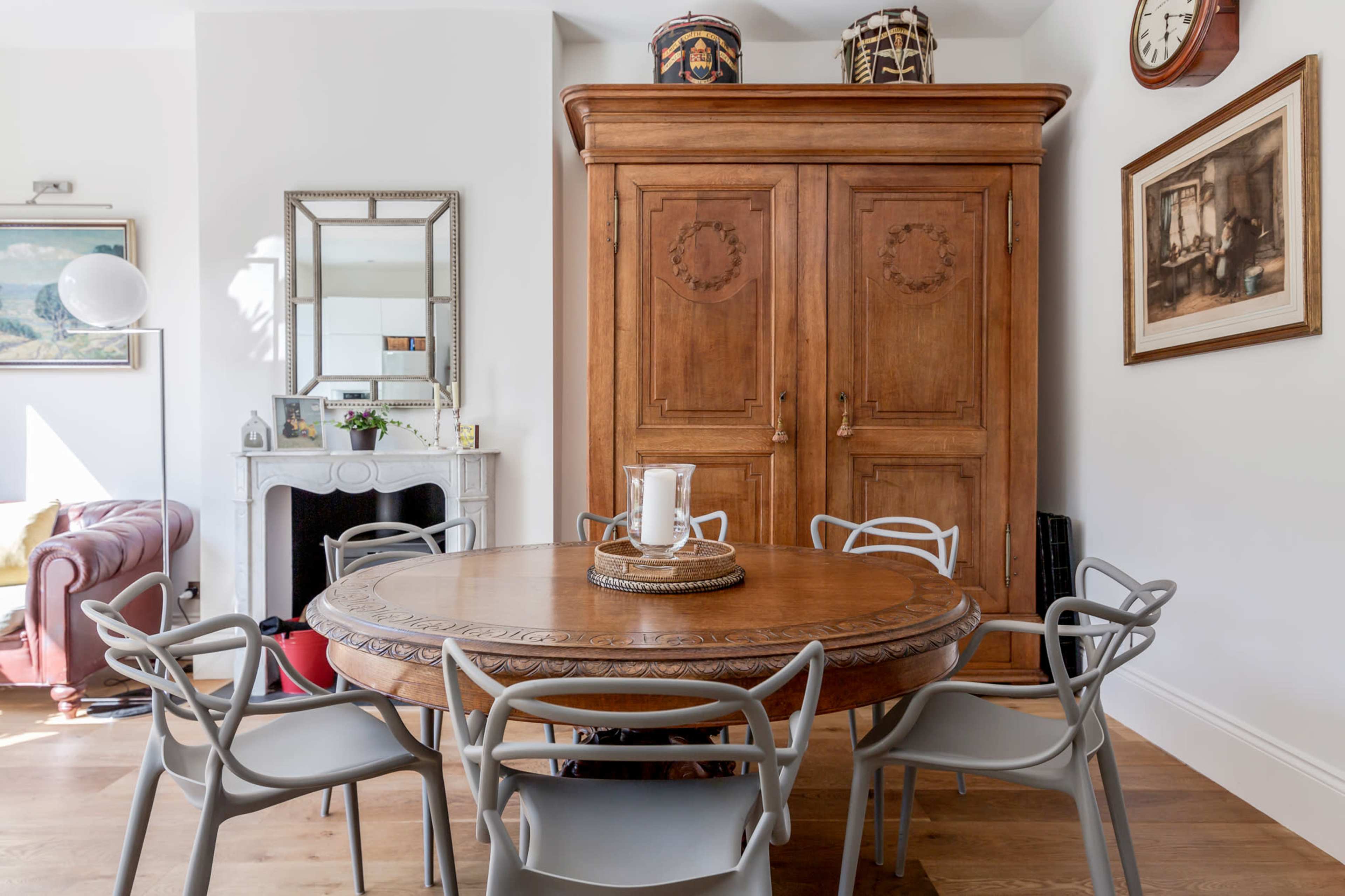 A round wooden dining table is surrounded by six chairs in a bright room featuring a large wooden cabinet and decorative wall art.