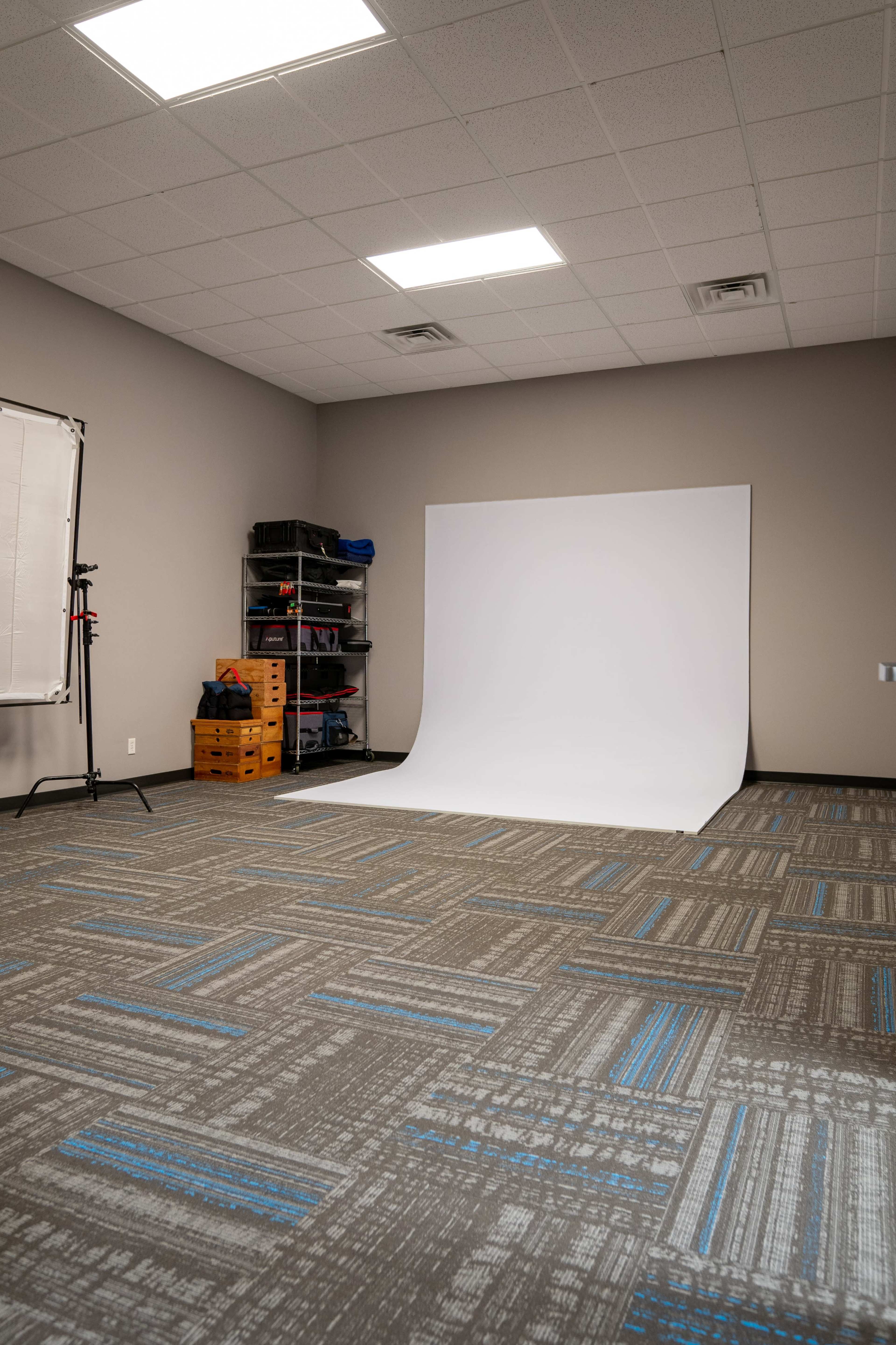 The image shows a photography studio with a white backdrop, storage shelves, and a carpeted floor with a patterned design.