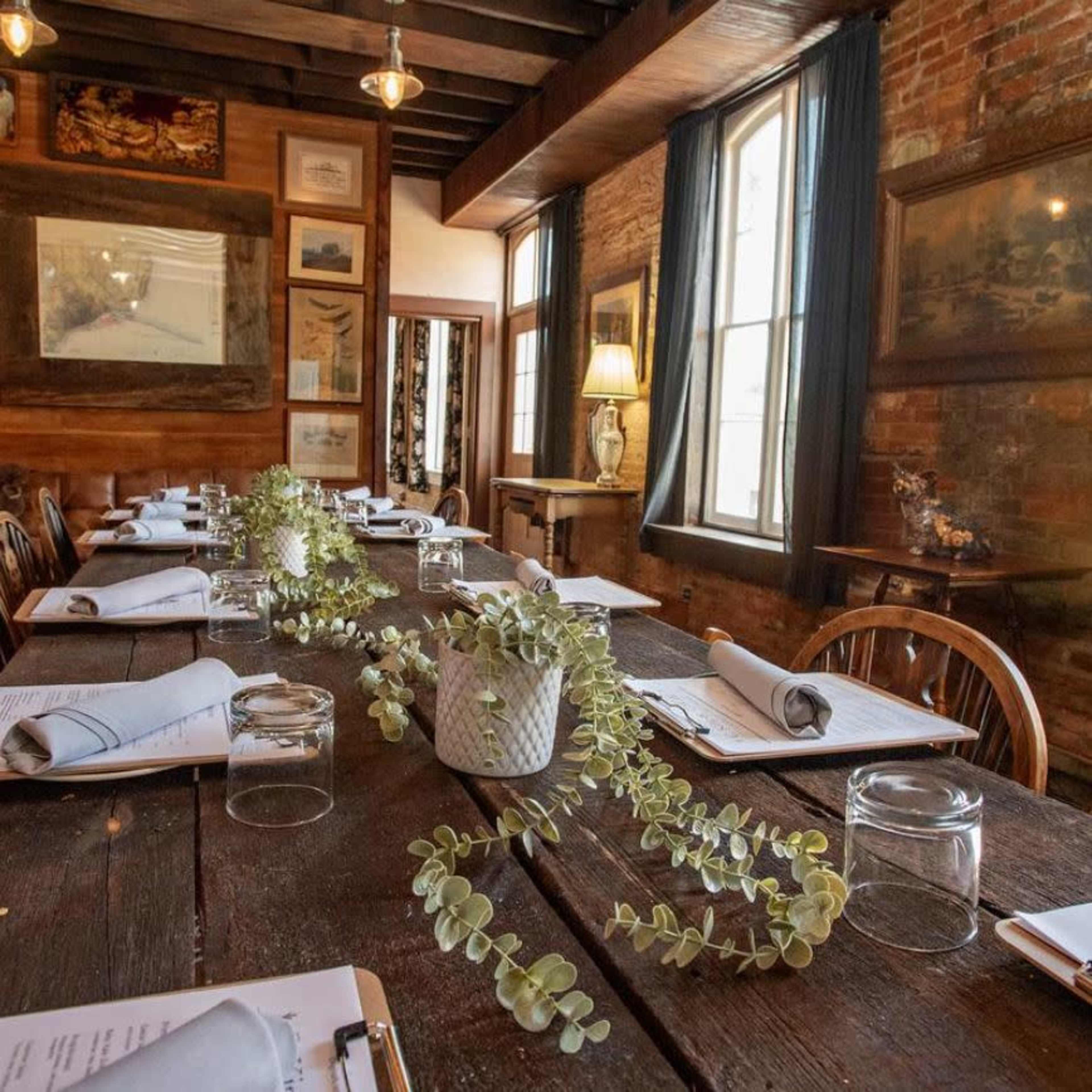 A long wooden table is set for a meal in a rustic room featuring exposed brick walls, large windows, and decorative artwork.