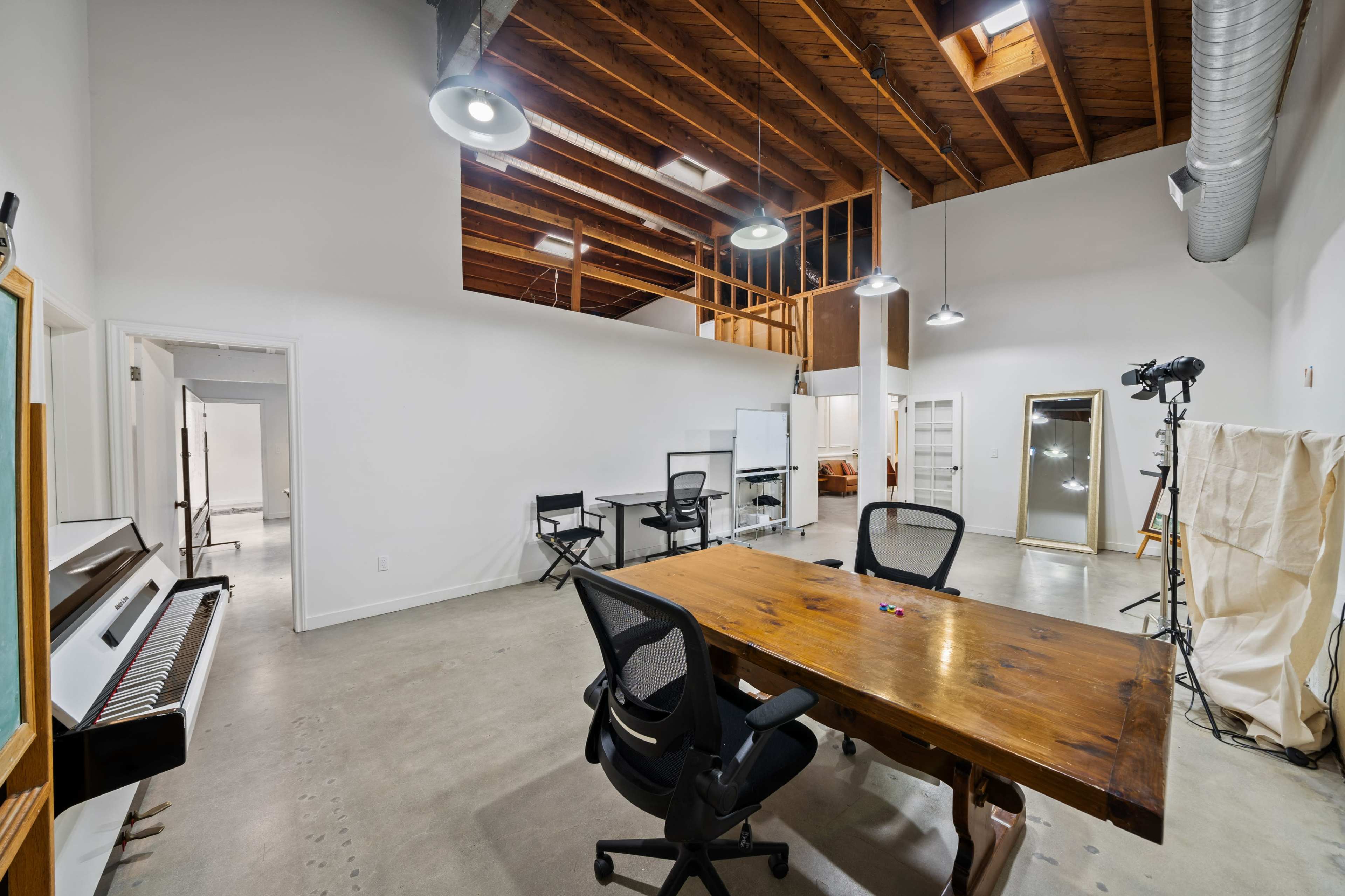 The image shows an open workspace with a wooden table, office chairs, a piano, and large windows illuminating the high ceiling with exposed beams.