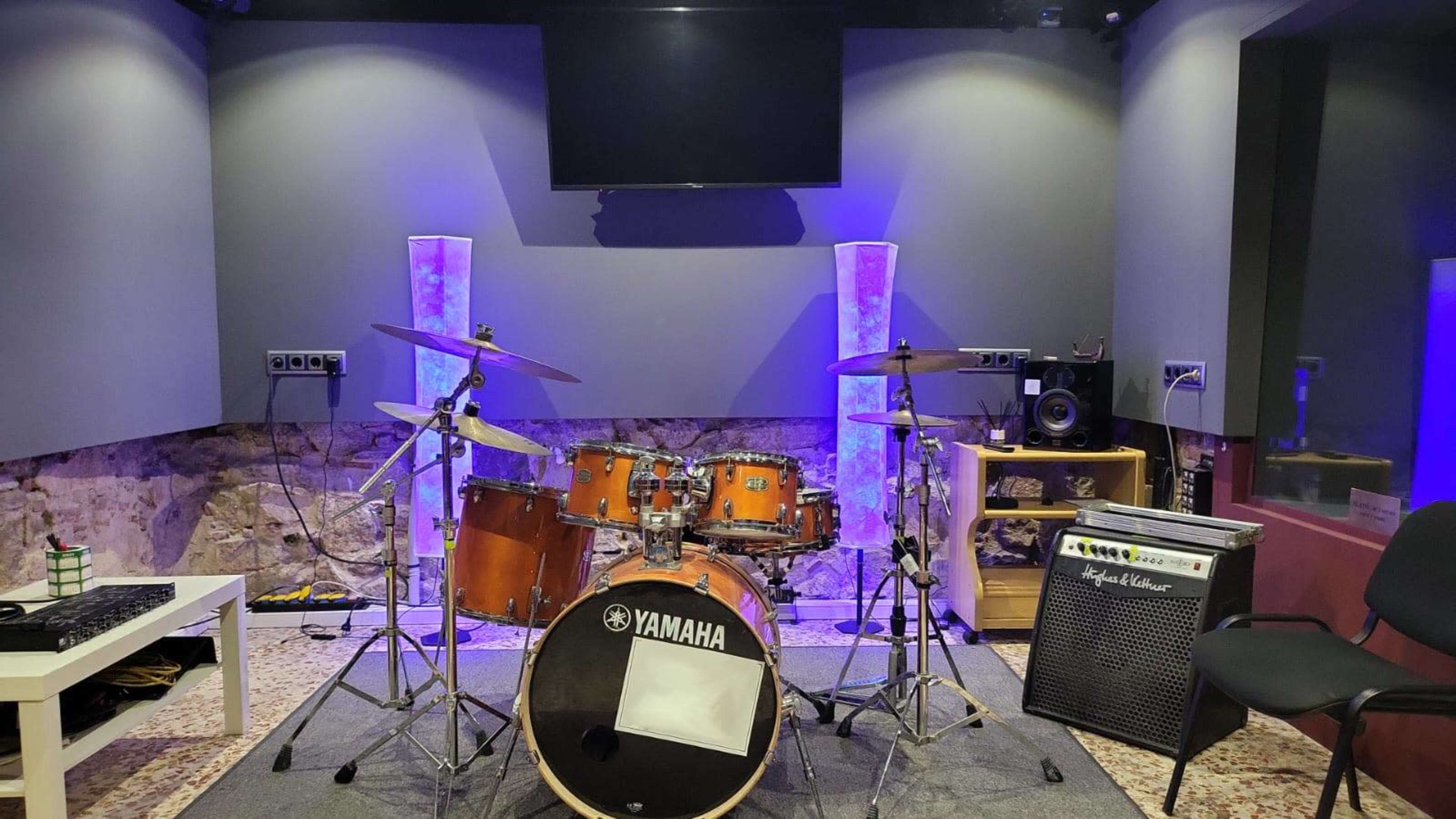 A music studio features a drum set, an amplifier, and a television screen, with purple ambient lighting and a stone wall backdrop.