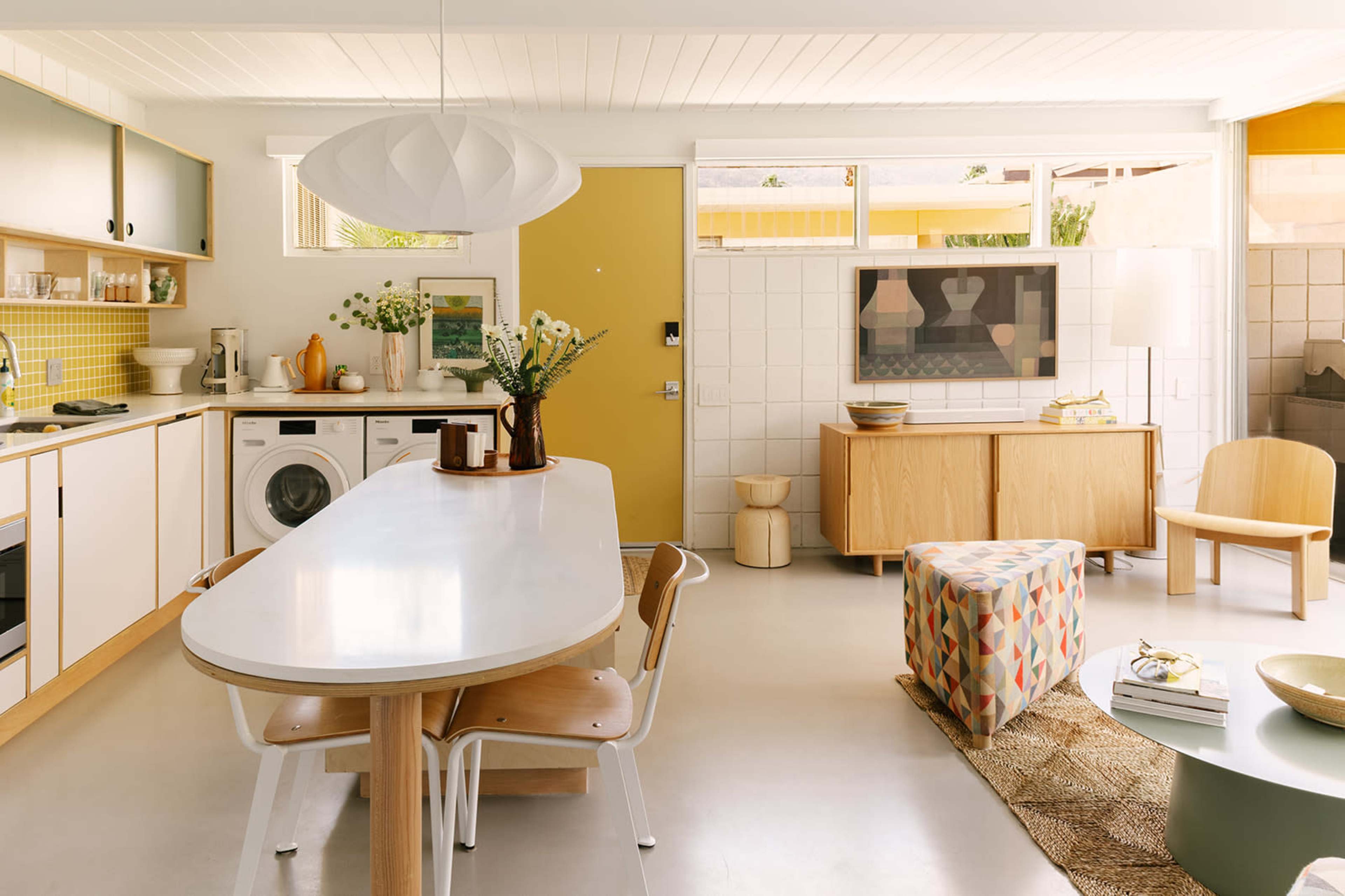 A modern kitchen and dining area features a long white table, light wood furniture, and large windows that allow natural light to enter.