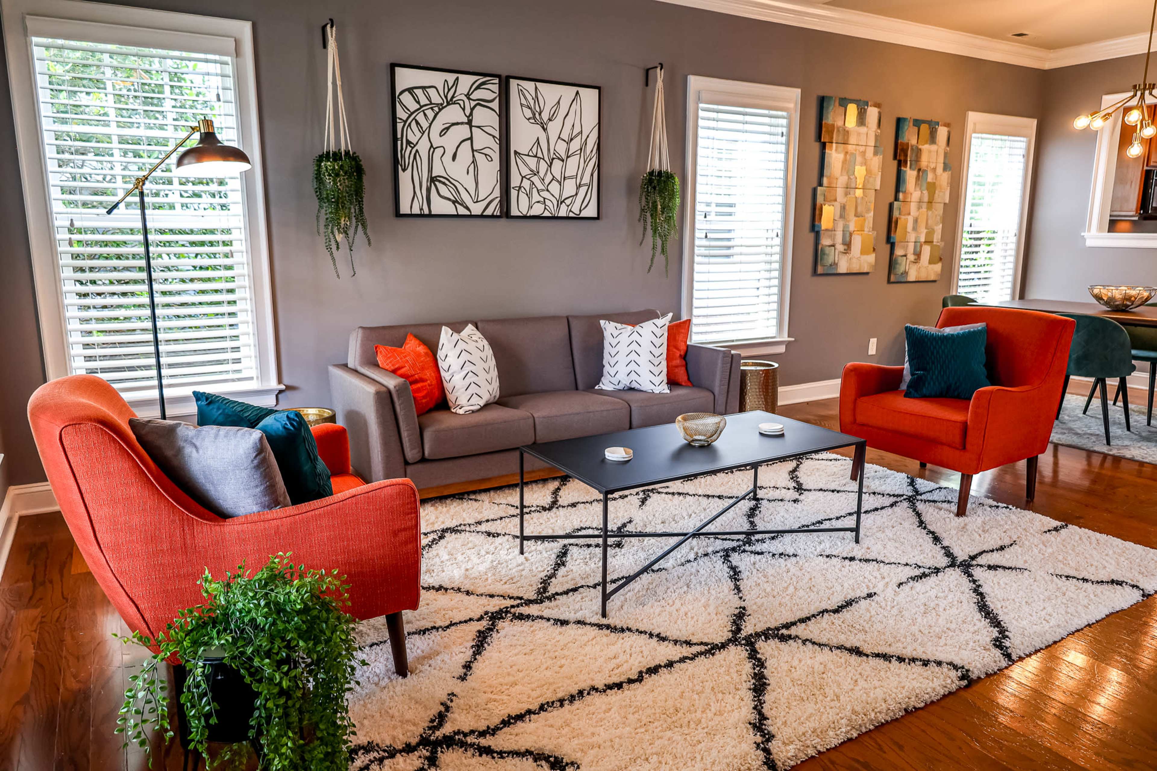 A modern living room with a gray couch and two orange armchairs arranged around a black coffee table on a patterned rug, with decorative artwork on the walls and natural light coming through the windows.