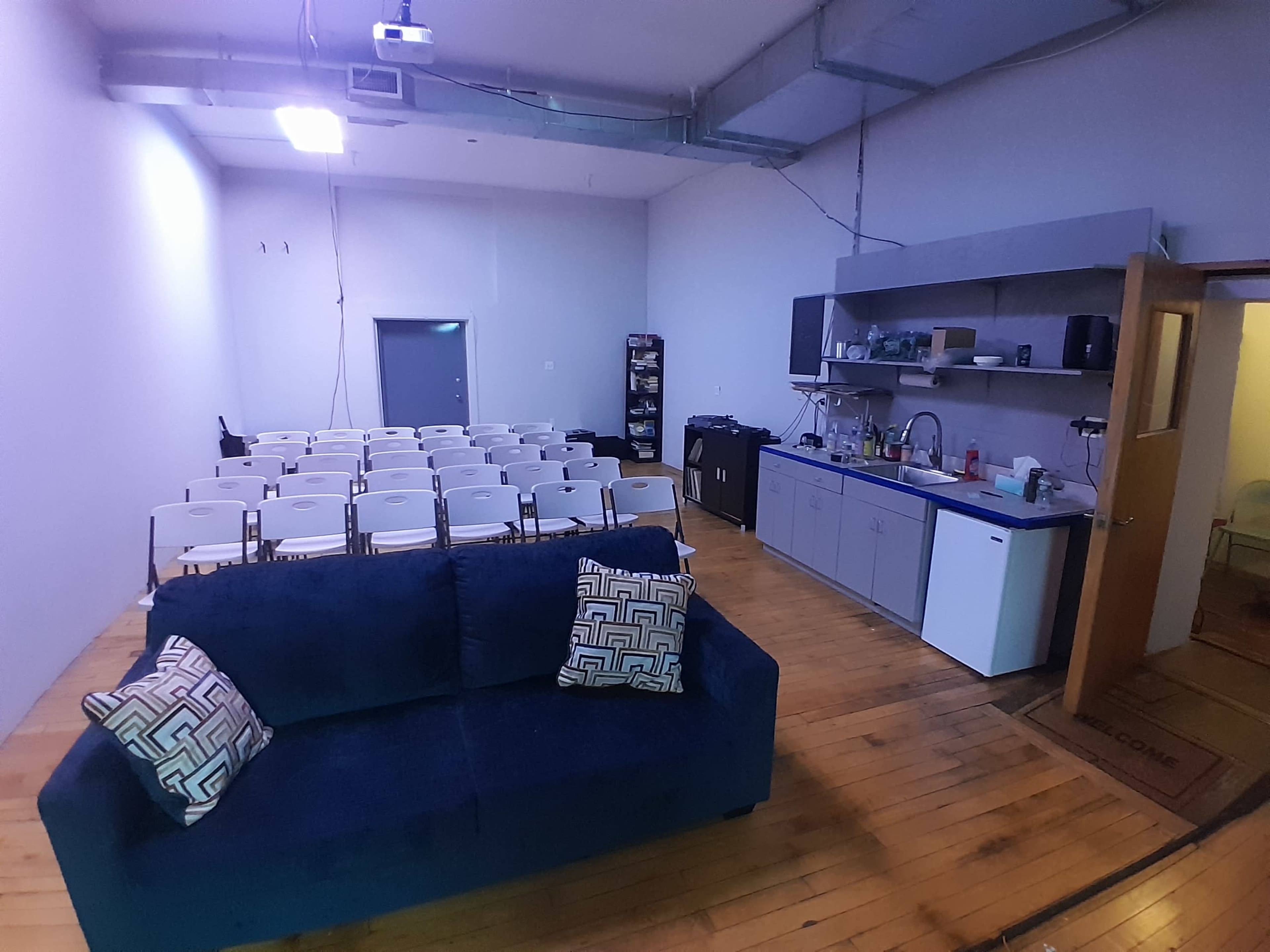 The image shows a spacious room with a blue couch in the foreground and rows of white chairs arranged for seating, along with a kitchenette area against the wall.