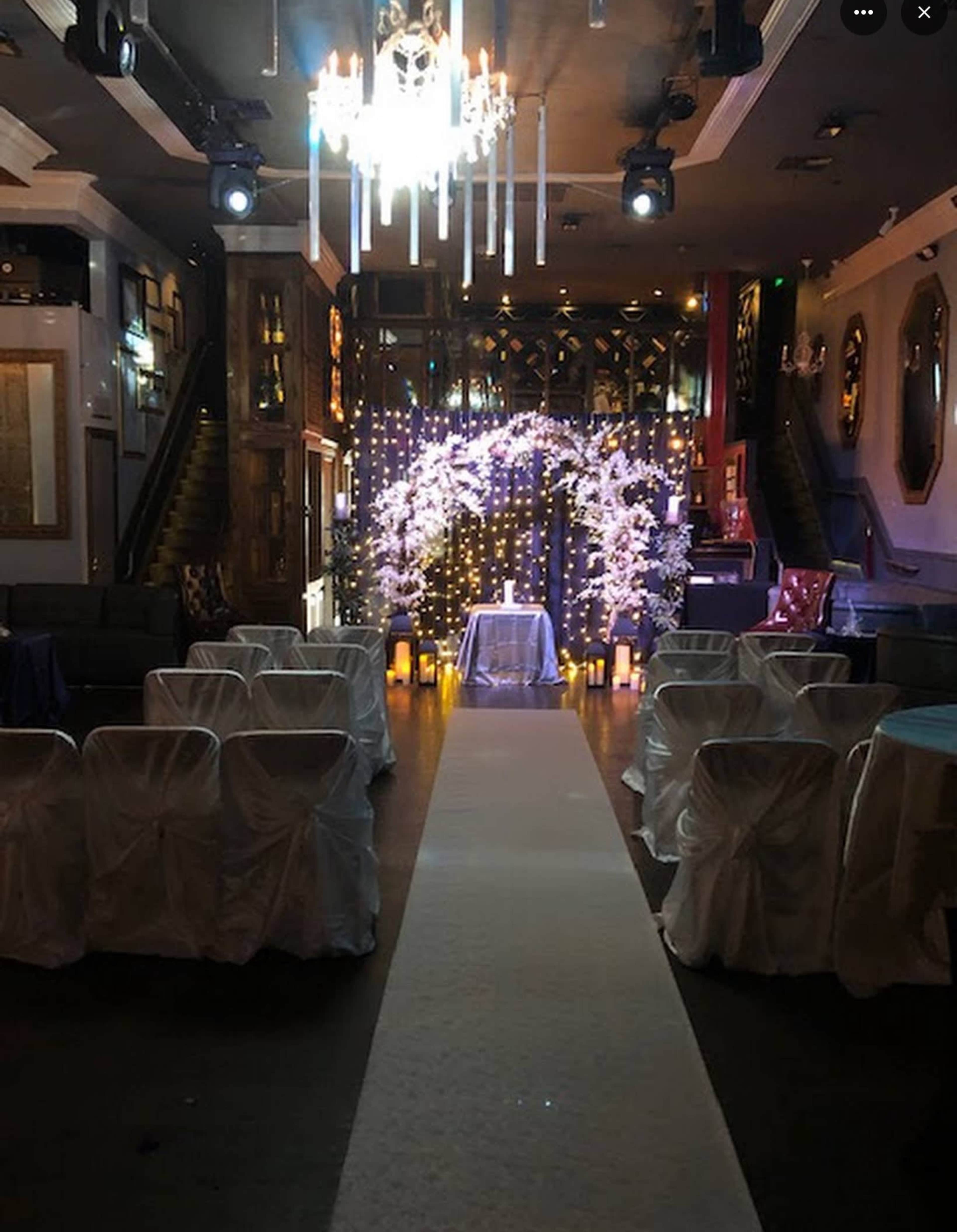 The image shows a decorated event space with an aisle leading to an arch adorned with lights and floral accents, surrounded by chairs draped in white fabric.