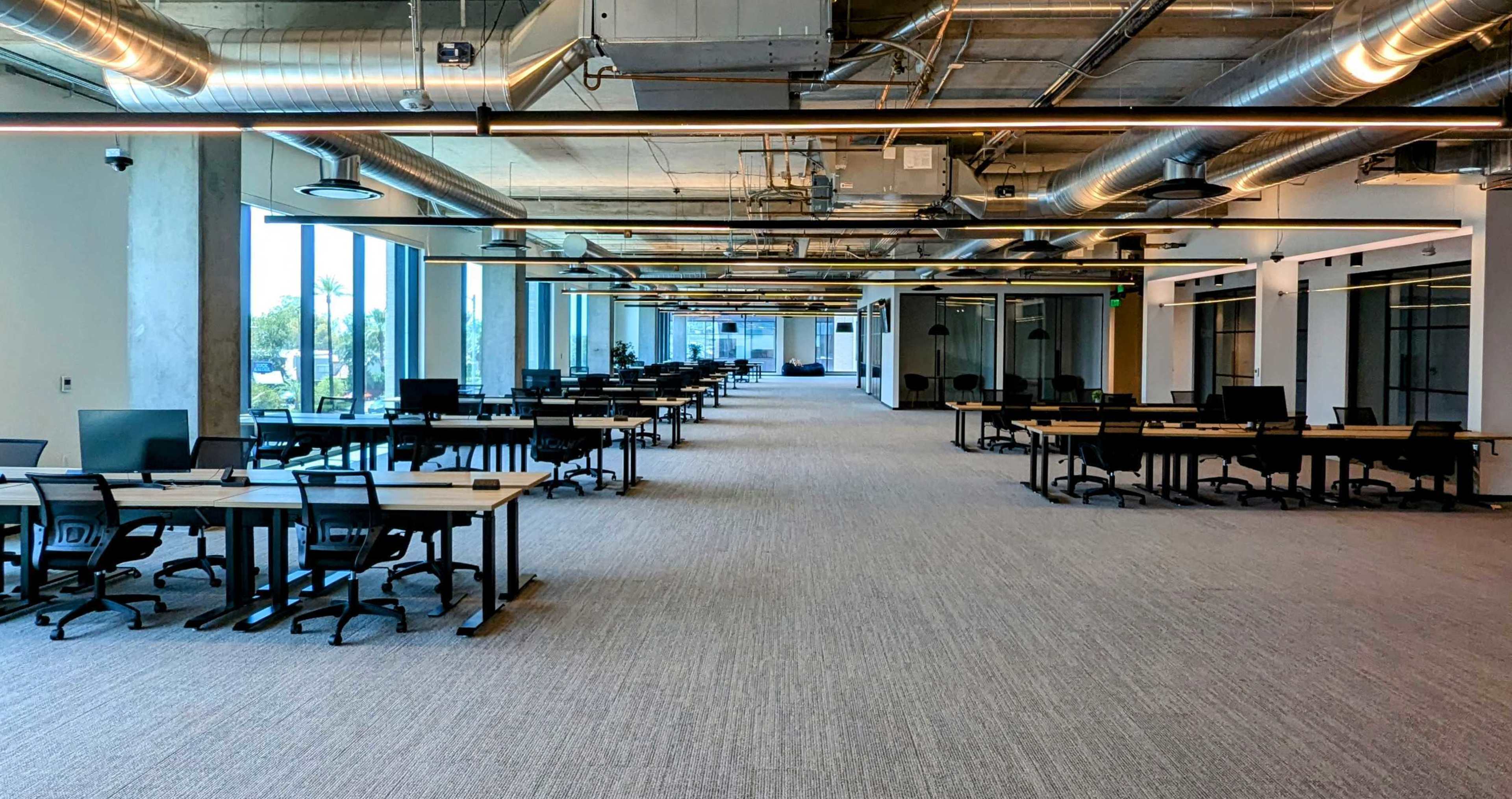 A spacious, modern office environment with rows of empty desks and large windows allowing natural light to enter.
