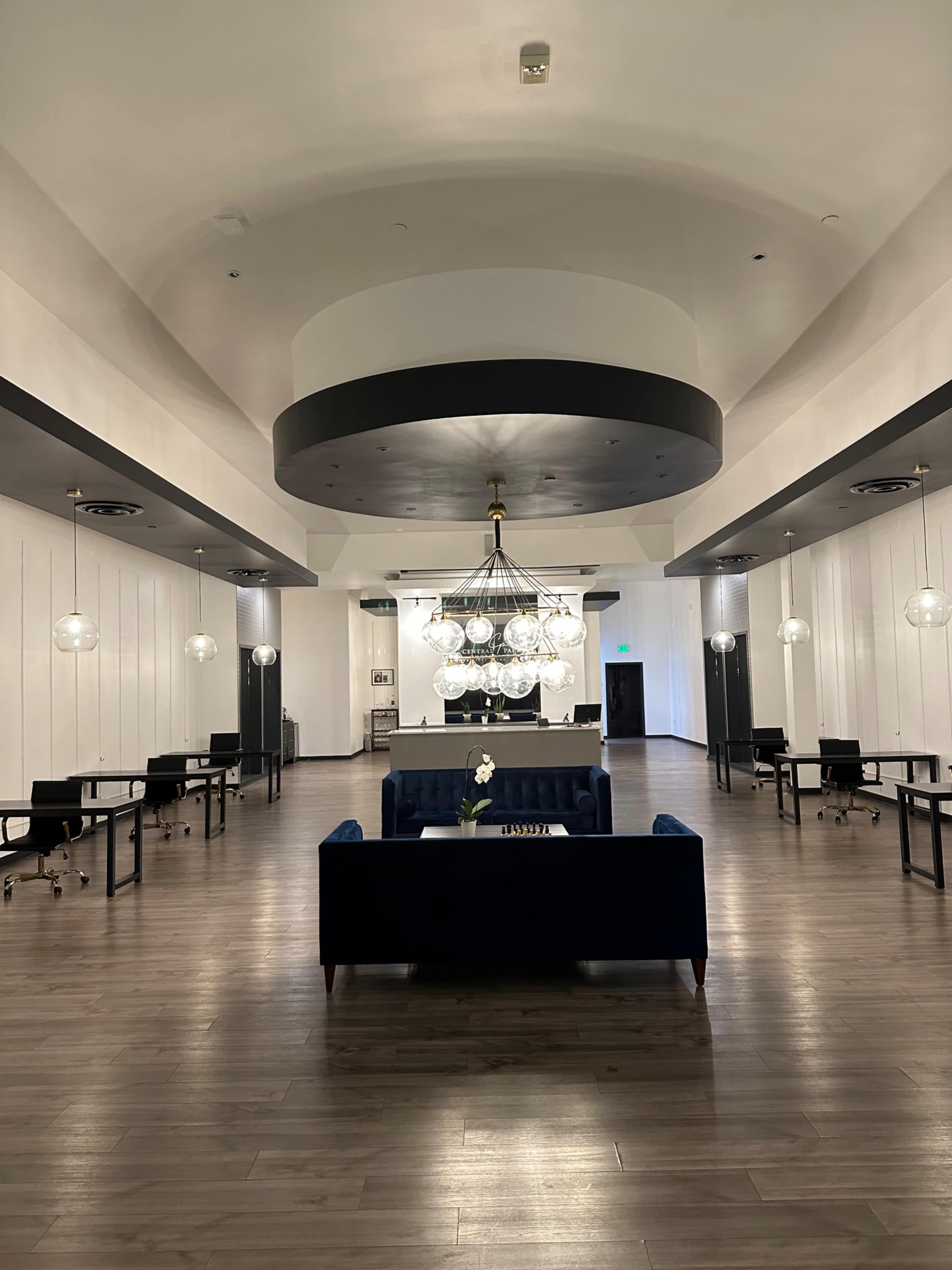 A spacious lobby area featuring a central chandelier, a blue sofa, and several desks arranged along the walls.