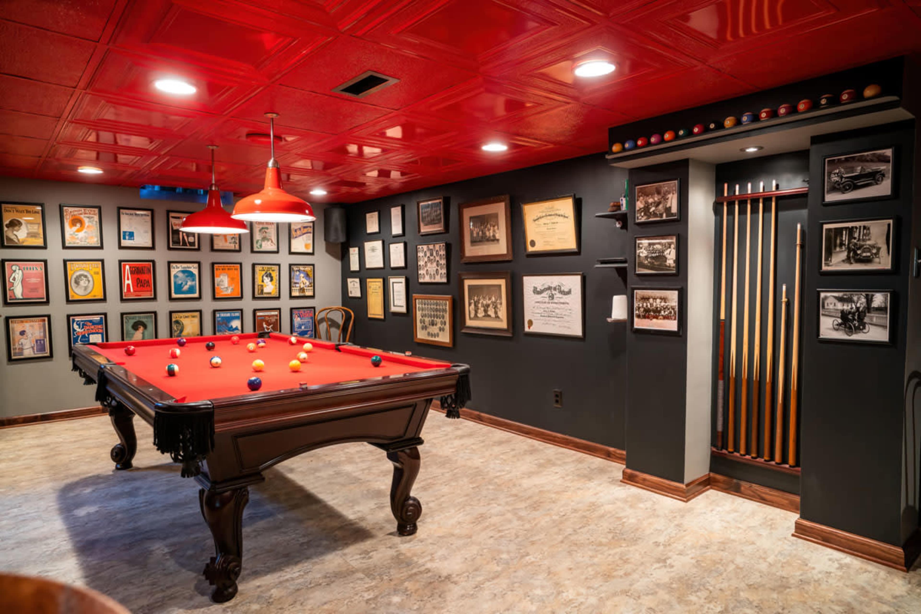 A billiard room featuring a red felt pool table surrounded by framed artwork and a wall-mounted rack for cue sticks.