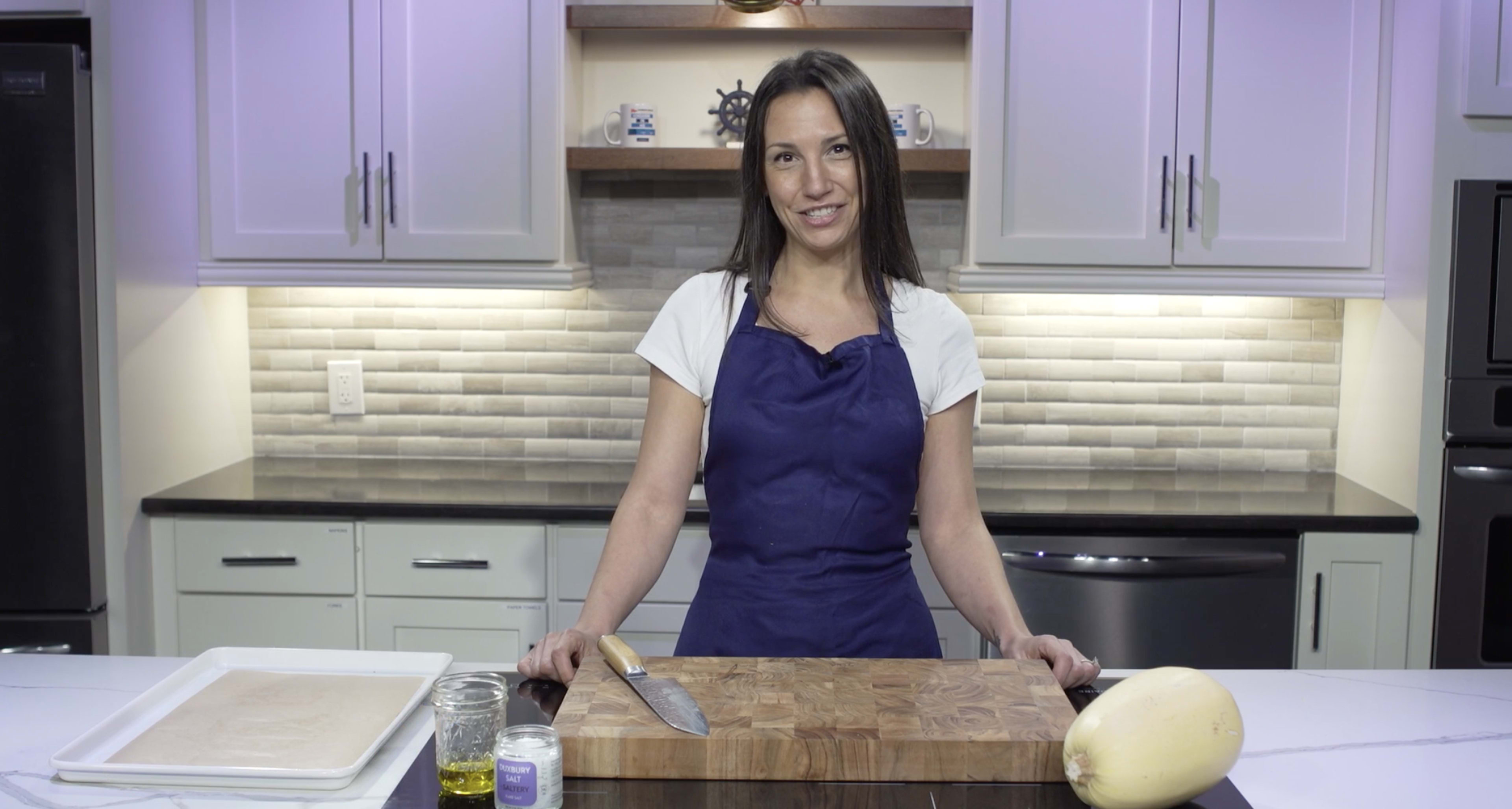A person wearing an apron stands behind a wooden cutting board in a kitchen, with a knife, a jar of oil, and a squash on the countertop.