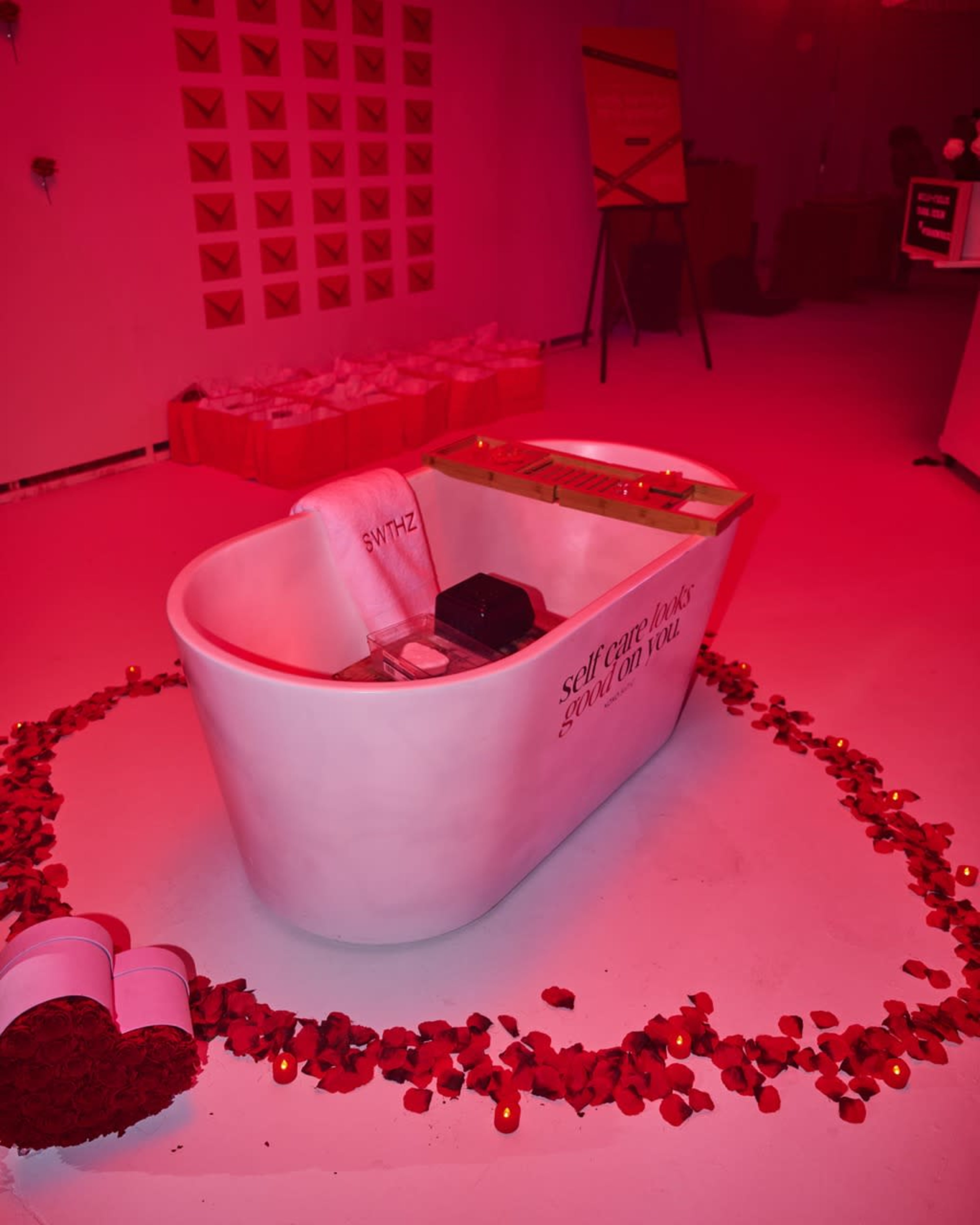 A white bathtub surrounded by rose petals and candles is the focal point in a dimly lit pink room.