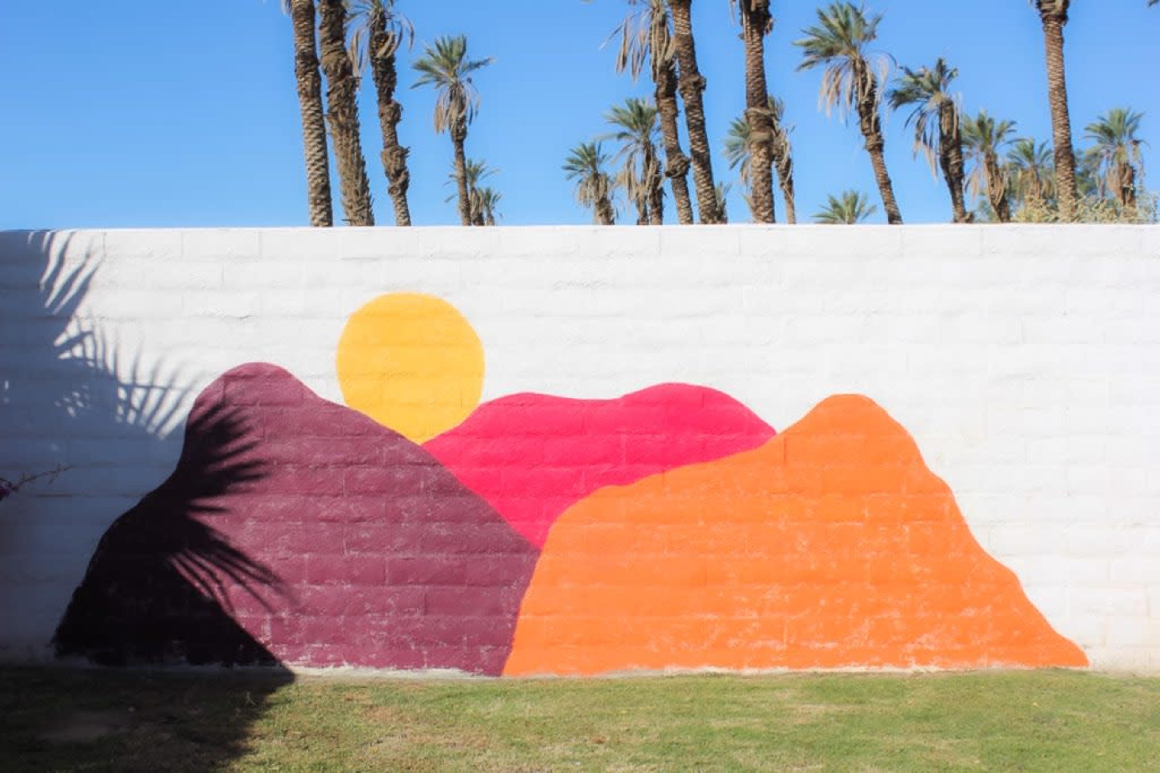 A colorful mural featuring stylized mountains and a sun is painted on a white wall, with palm trees visible in the background.