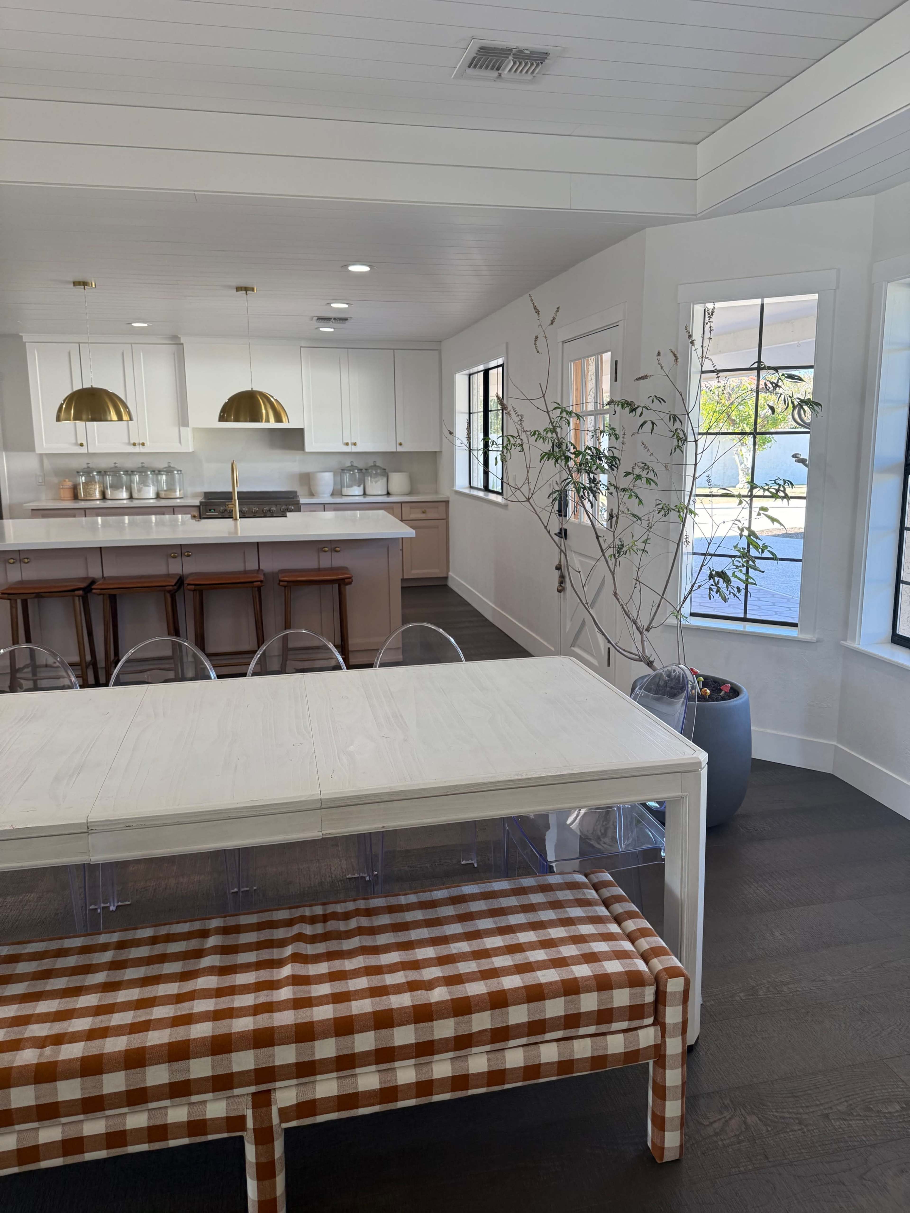 The image depicts a modern kitchen and dining area with a clear, light-colored dining table, transparent chairs, and a checkered bench, surrounded by large windows.