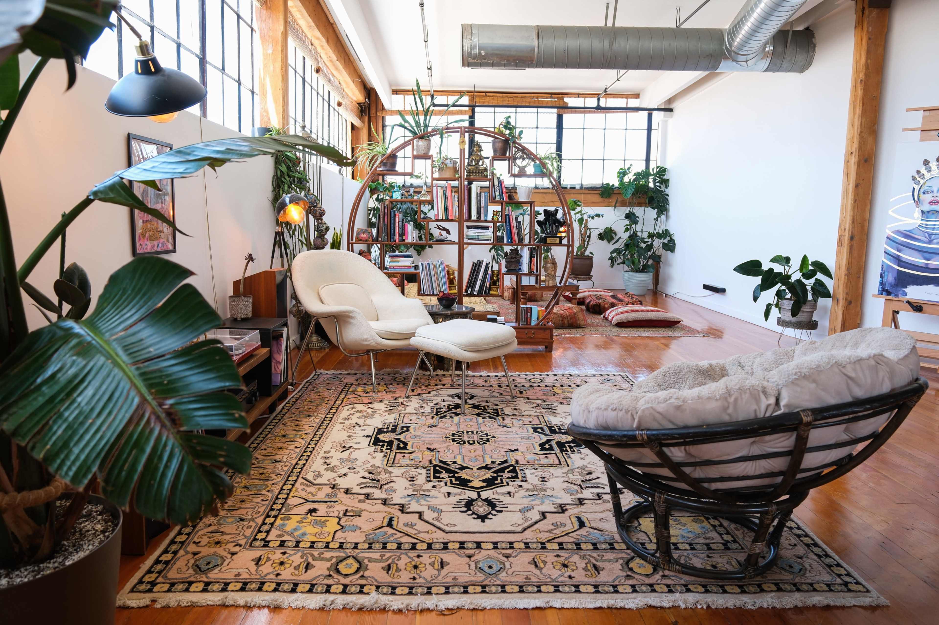 The image shows a cozy interior space featuring a plush chair, a white lounge chair, a large bookshelf filled with books, and several indoor plants.