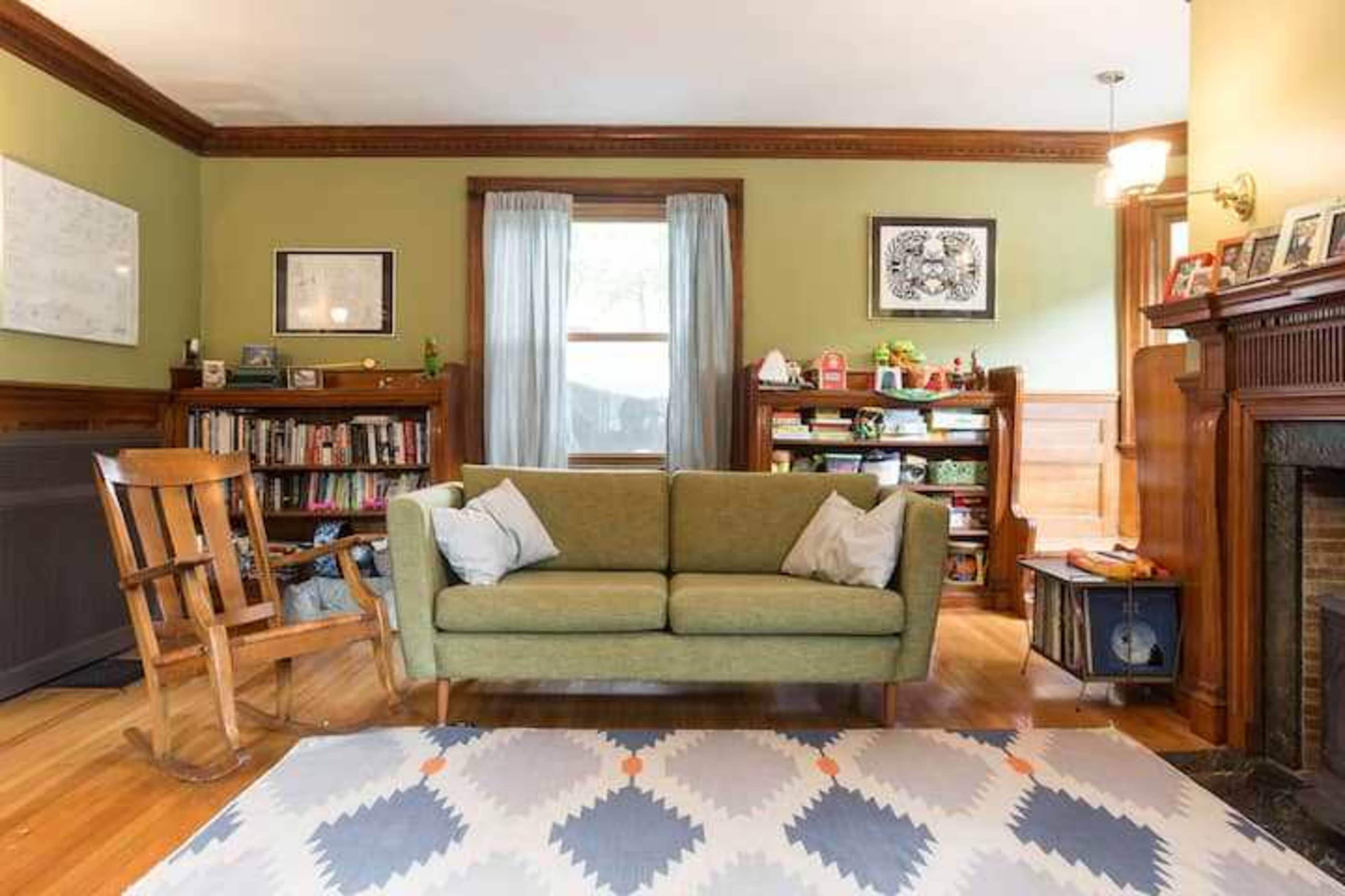 The living room features a green sofa, a wooden rocking chair, a bookshelf with various items, and a fireplace with a mantel, all set against a backdrop of light green walls and wooden trim.