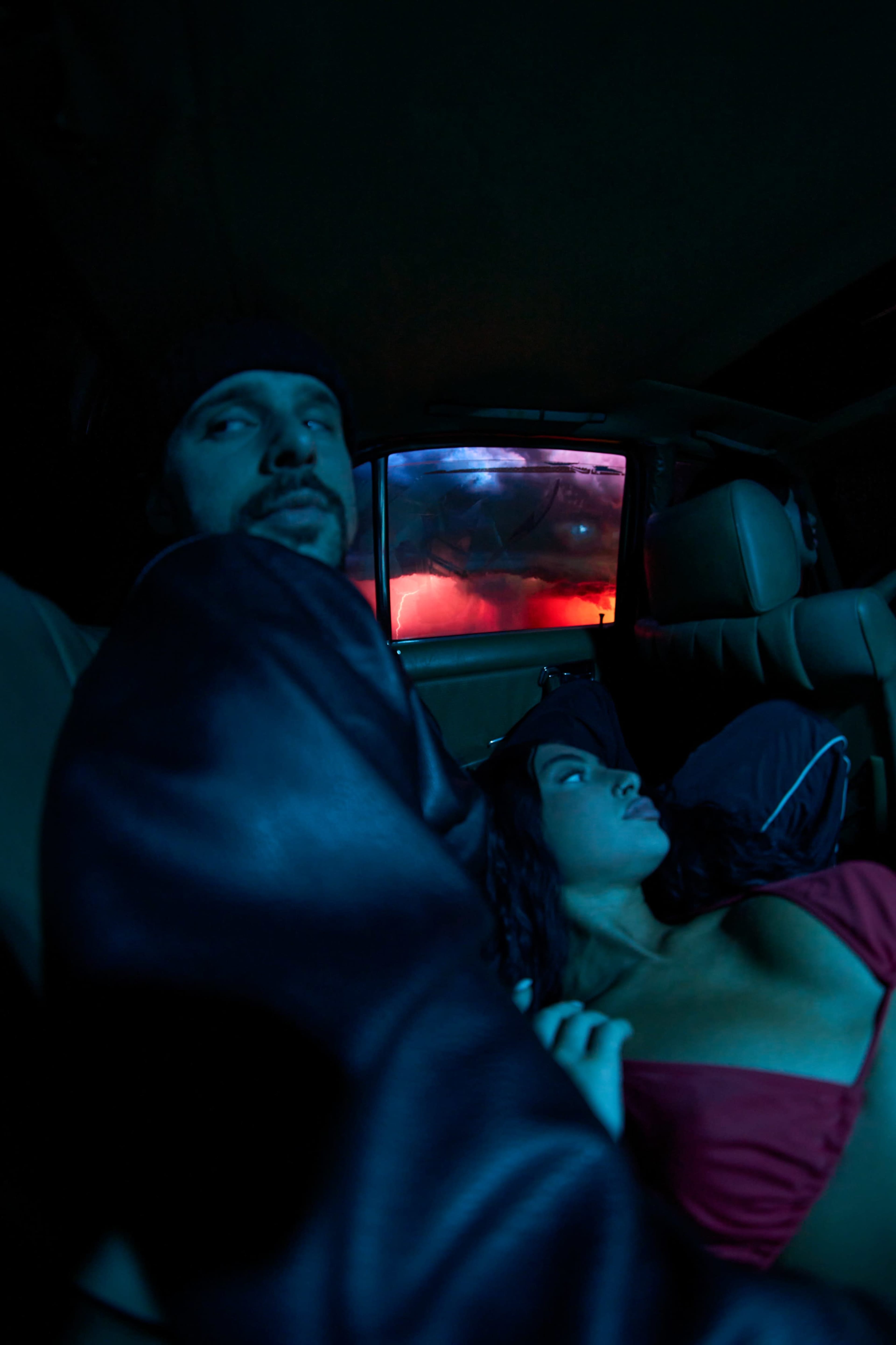 A man and woman are sitting in the backseat of a car, with blue and red lights illuminating the scene from outside.