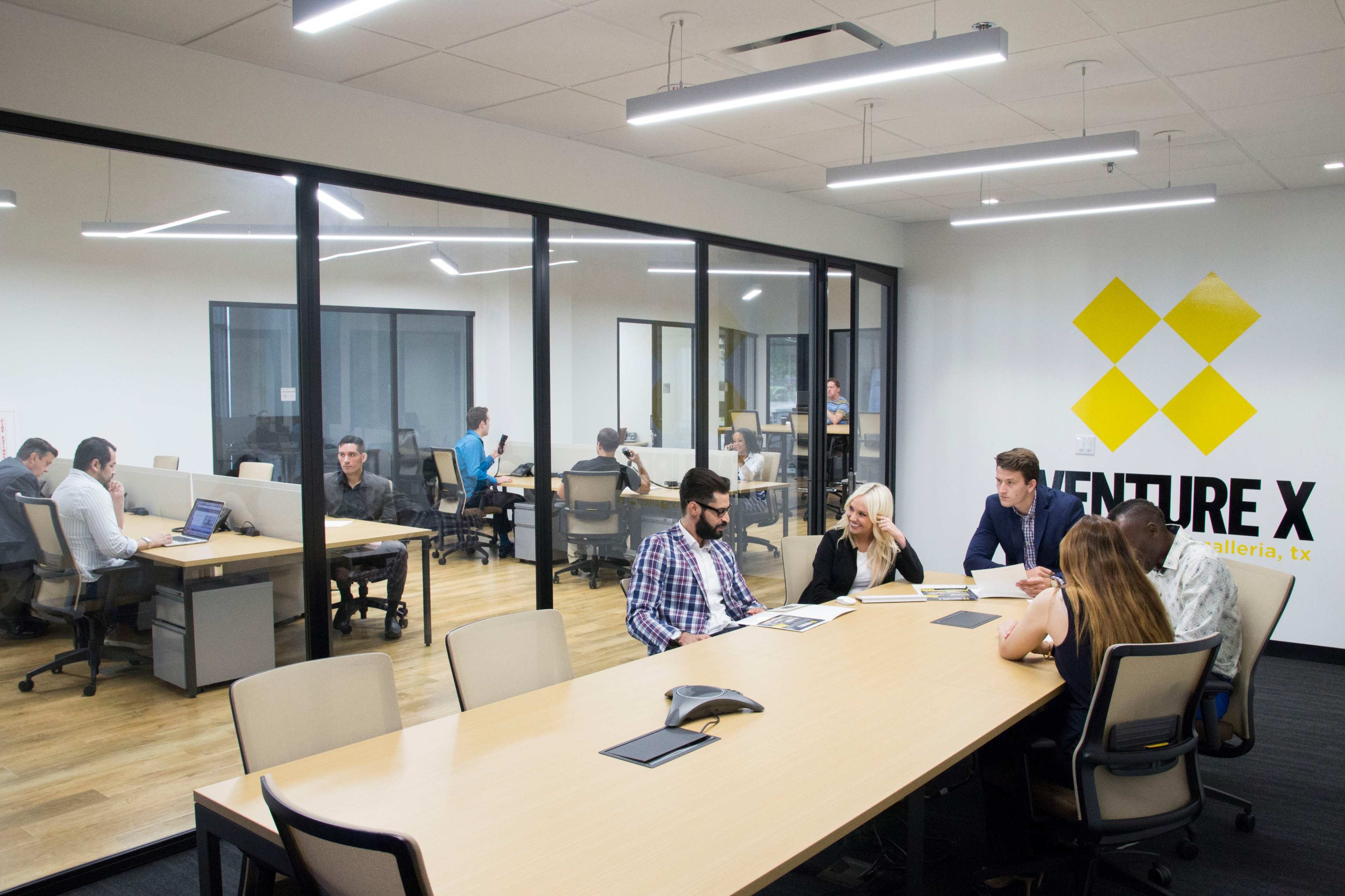 A group of professionals collaborates around a conference table in a modern office space, while others work at individual desks in the background.