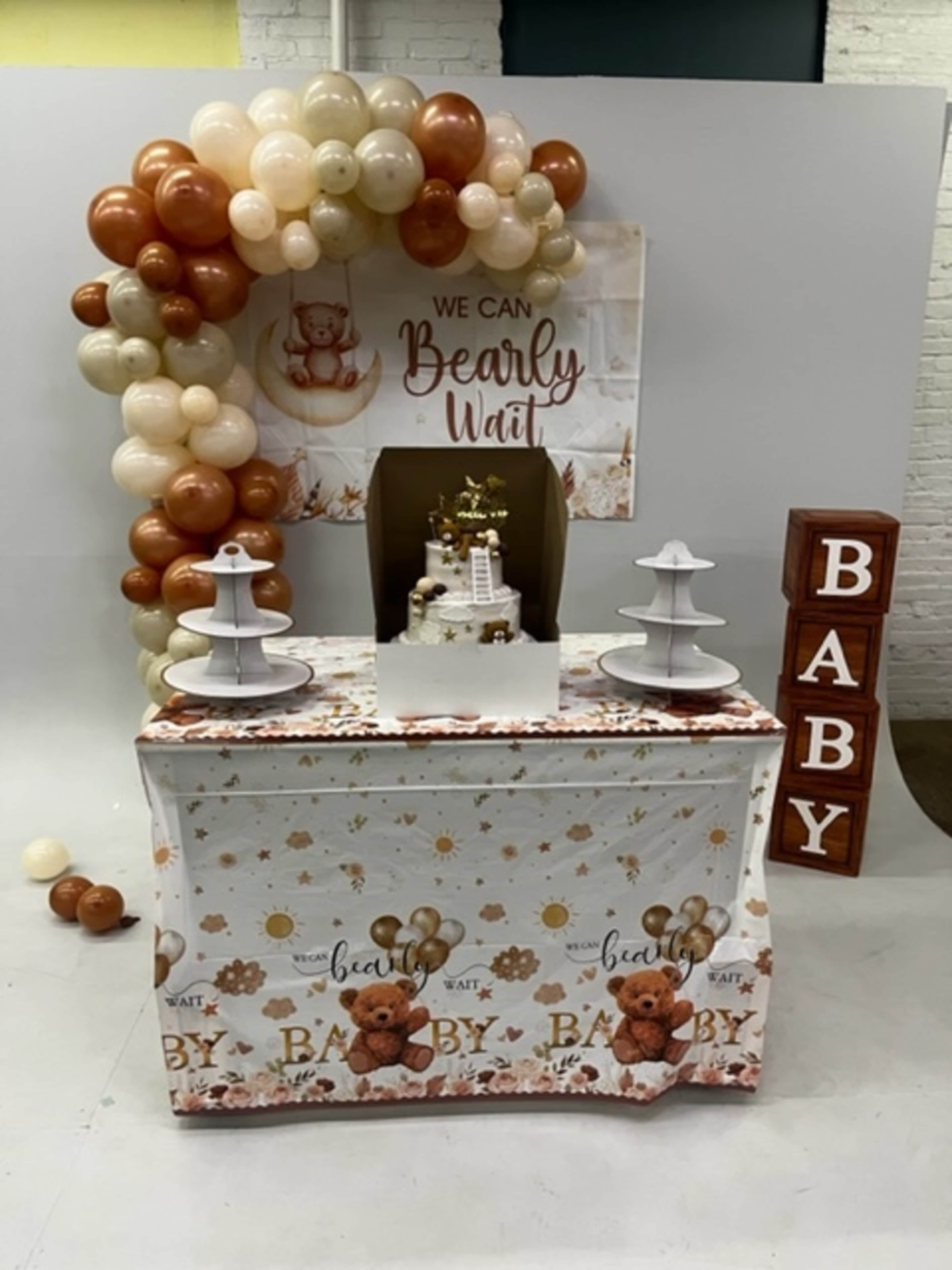 The image features a decorated table for a baby shower, complete with a cake on a stand, themed backdrop, and a display of balloons in earthy tones.