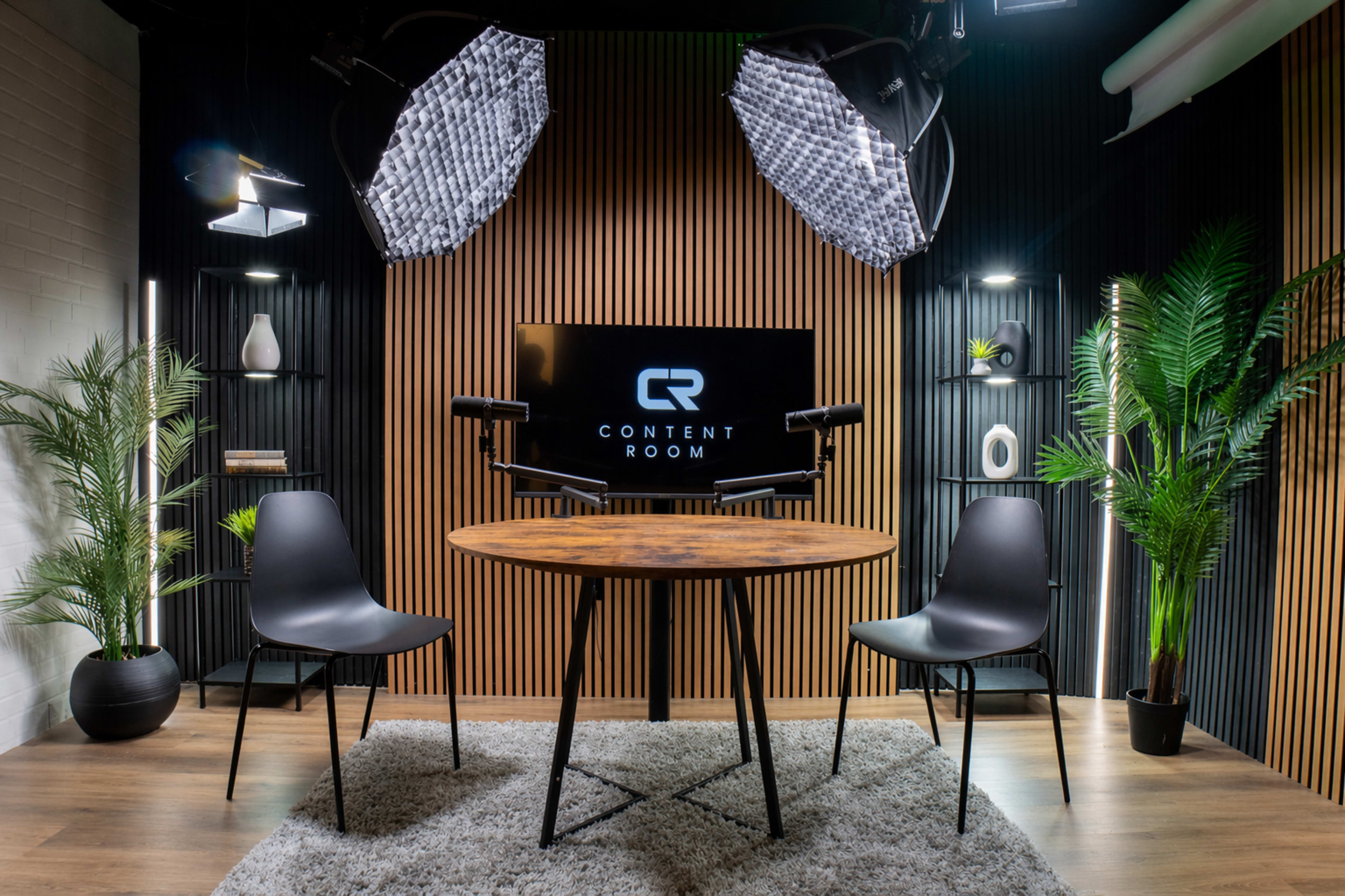 The image shows a modern content creation room equipped with a round wooden table, two black chairs, and a backdrop featuring the words "CONTENT ROOM" on a screen, surrounded by plants and studio lighting.