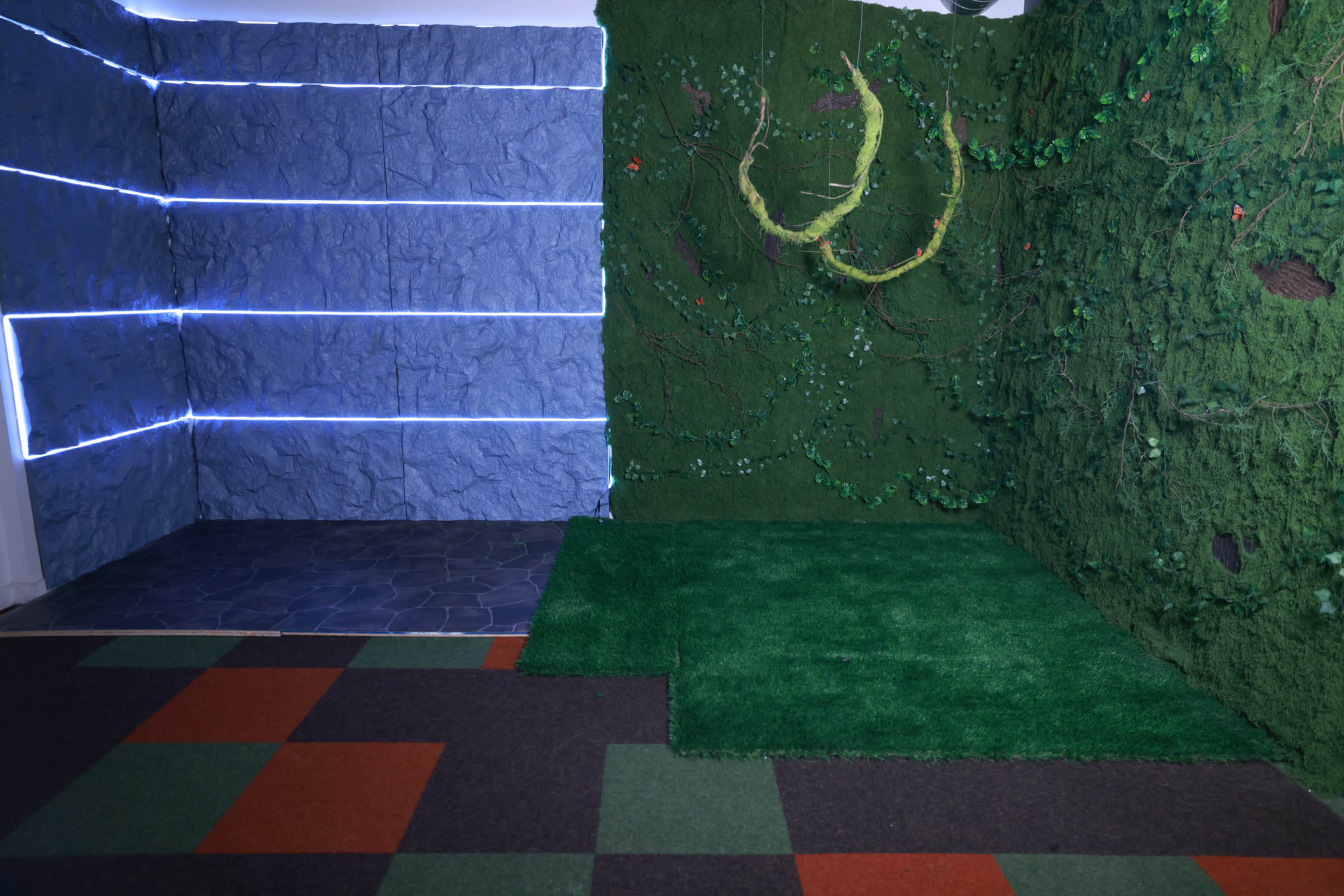 The image shows a room featuring a textured blue wall on one side and a green, leafy wall with vines on the other, along with a dark stone floor and a section of artificial grass.