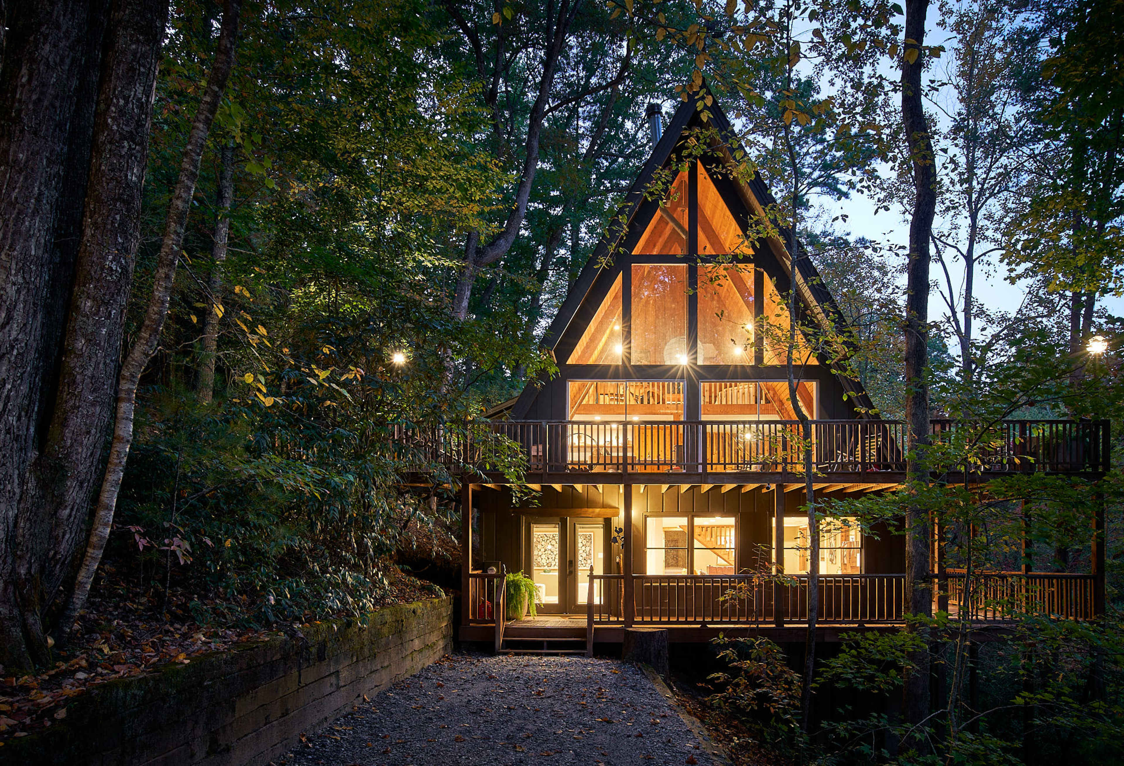 A two-story A-frame cabin with large windows is situated among trees, illuminated at dusk.