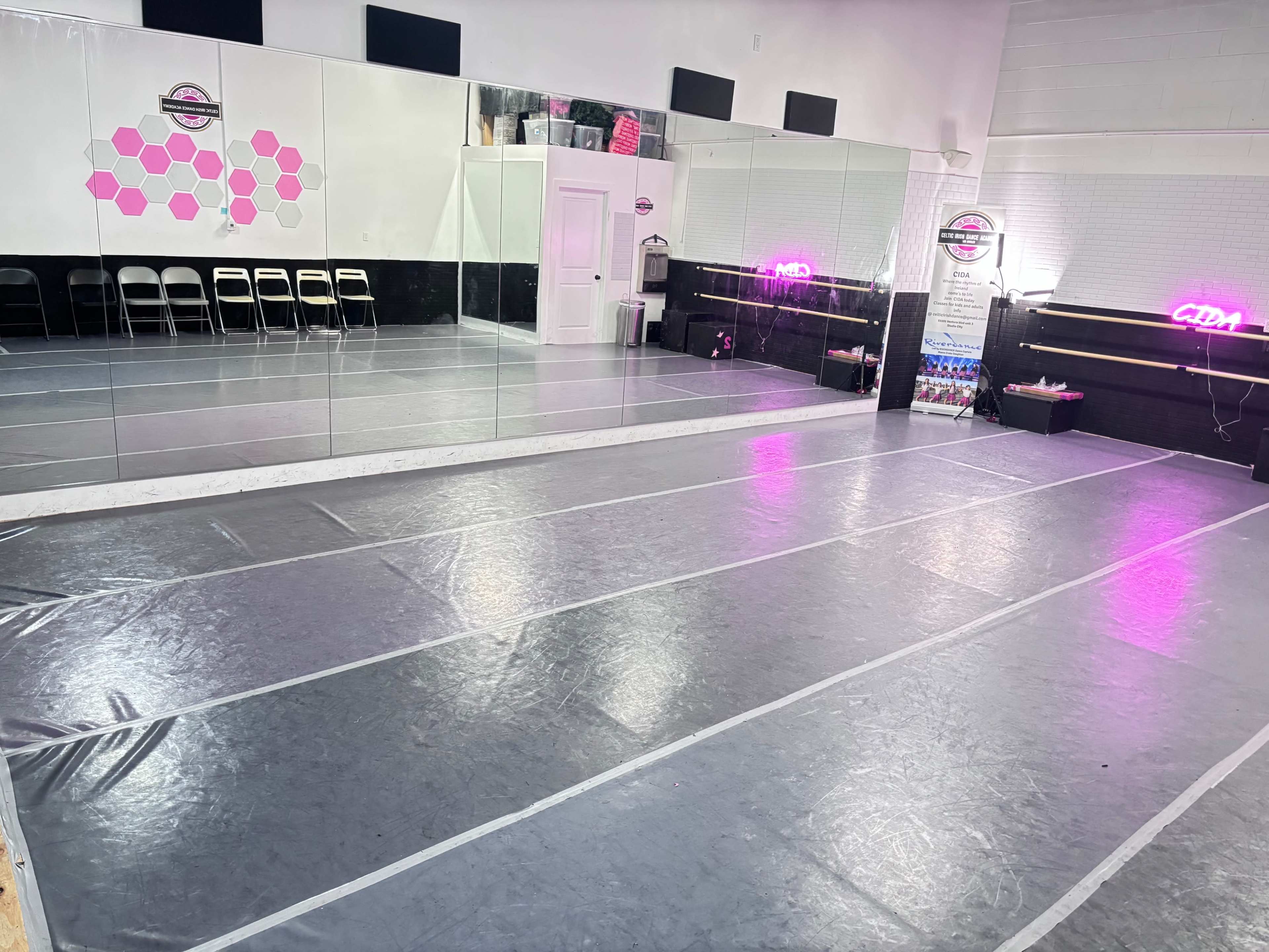 The image shows a dance studio with a mirrored wall and a shiny floor, featuring chairs along the back and neon lighting.