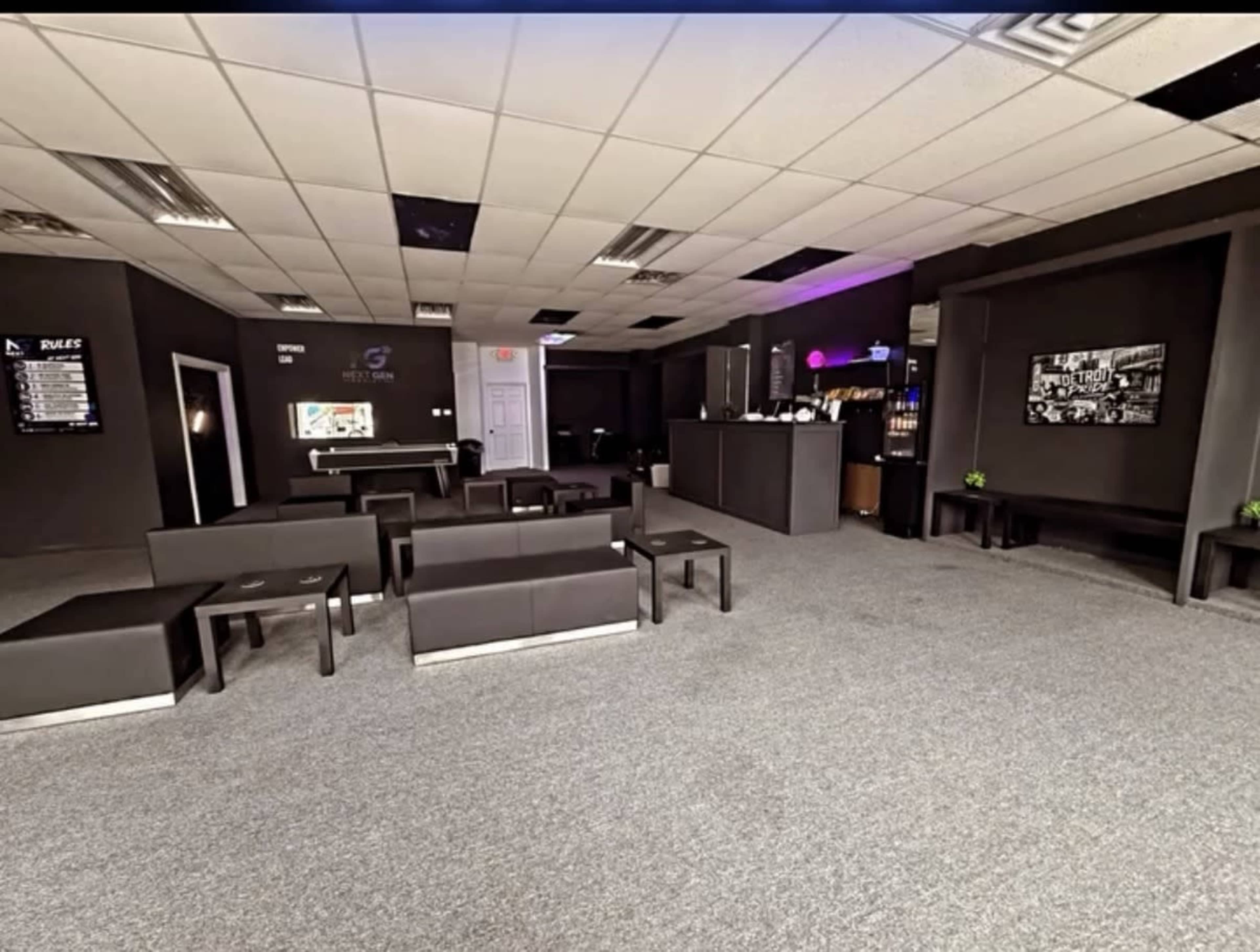 The image shows a spacious lounge area with several black couches, a reception desk, and a bar counter in a dimly lit room with gray carpet and dark walls.