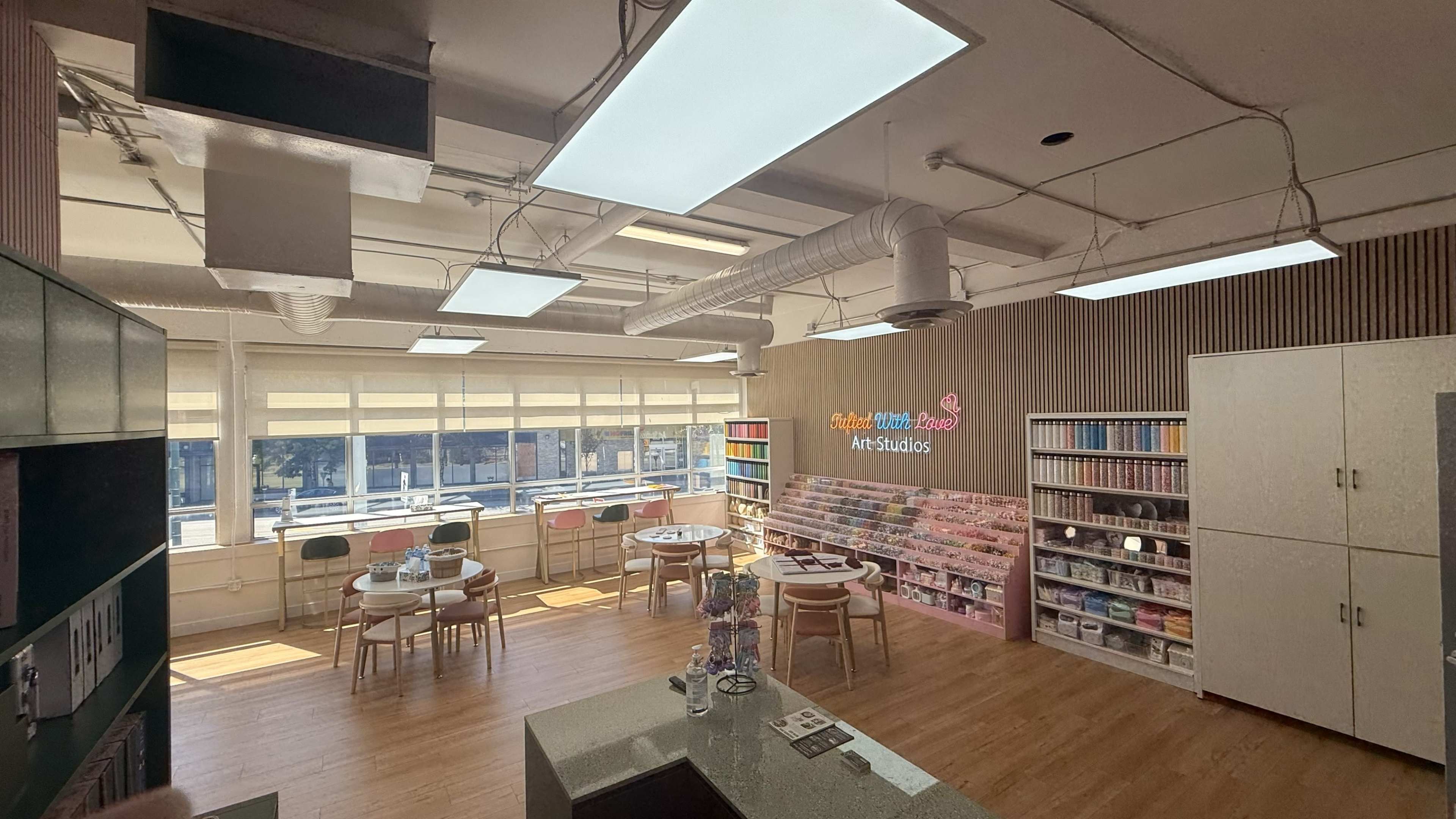 The image shows a brightly lit art studio with tables, shelves of art supplies, and large windows offering natural light.