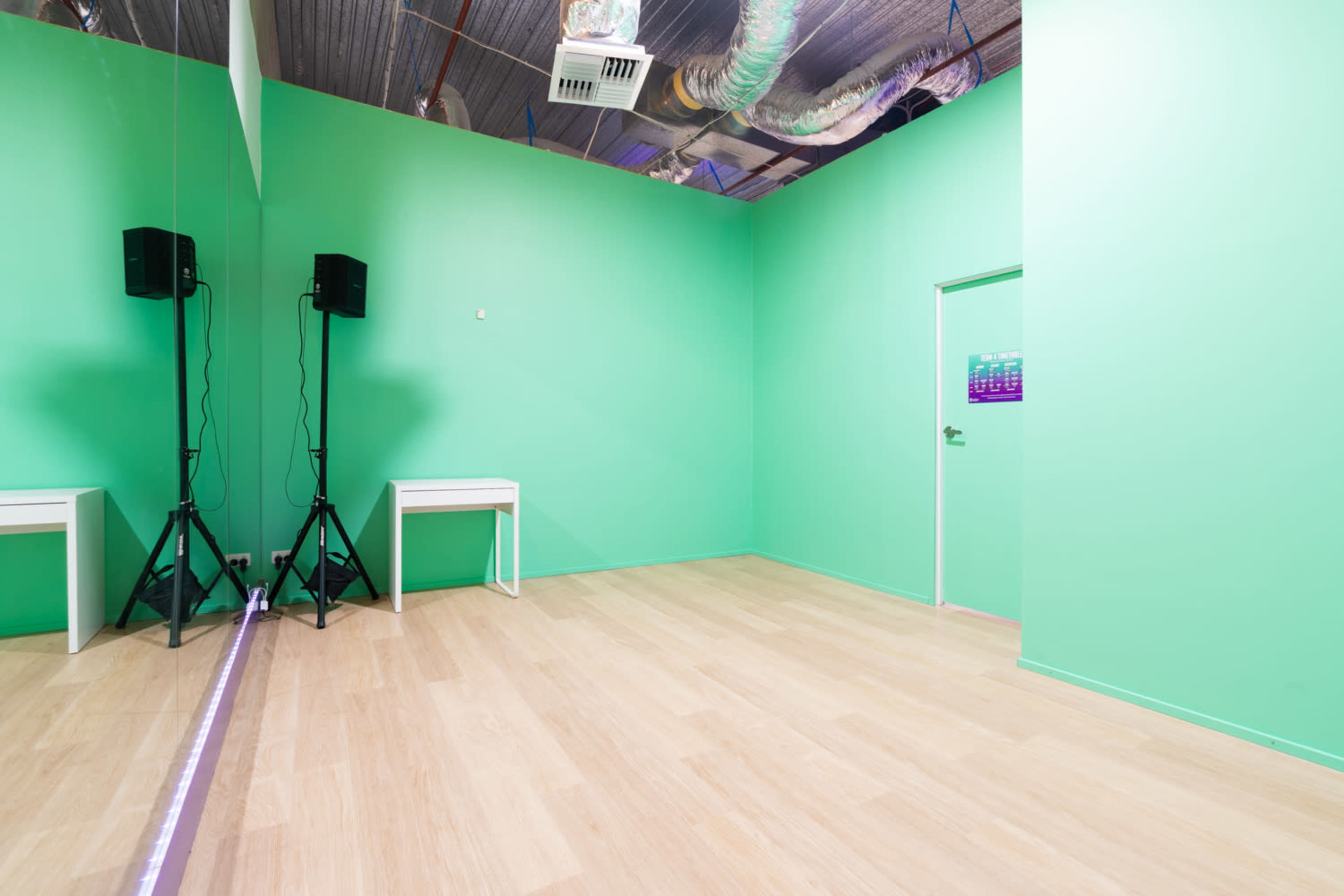 A room with green walls, wooden flooring, a mirrored wall, two speakers on stands, and a door.