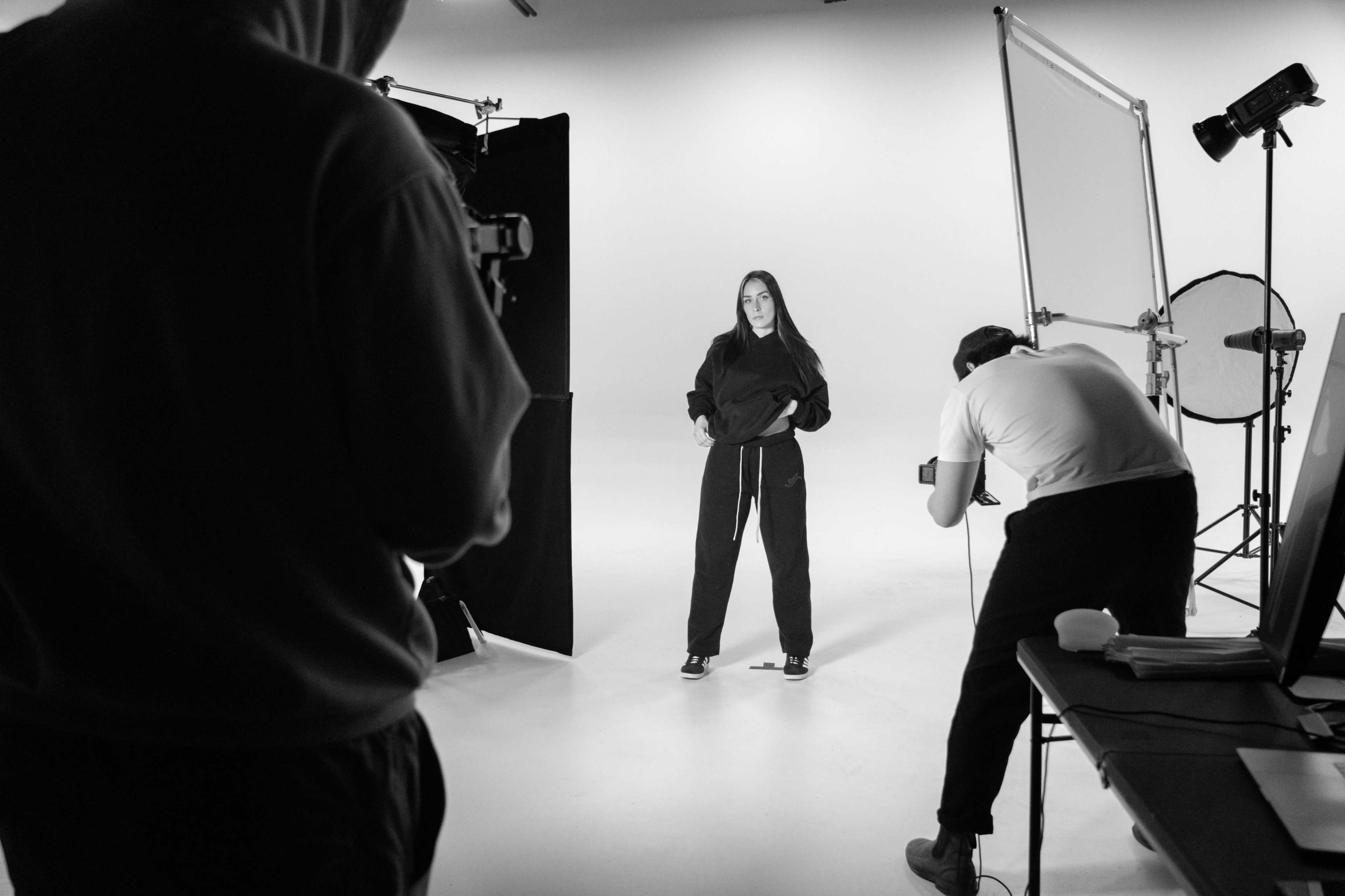 A person in a hooded outfit poses for a photo while another individual sets up a camera in a studio.