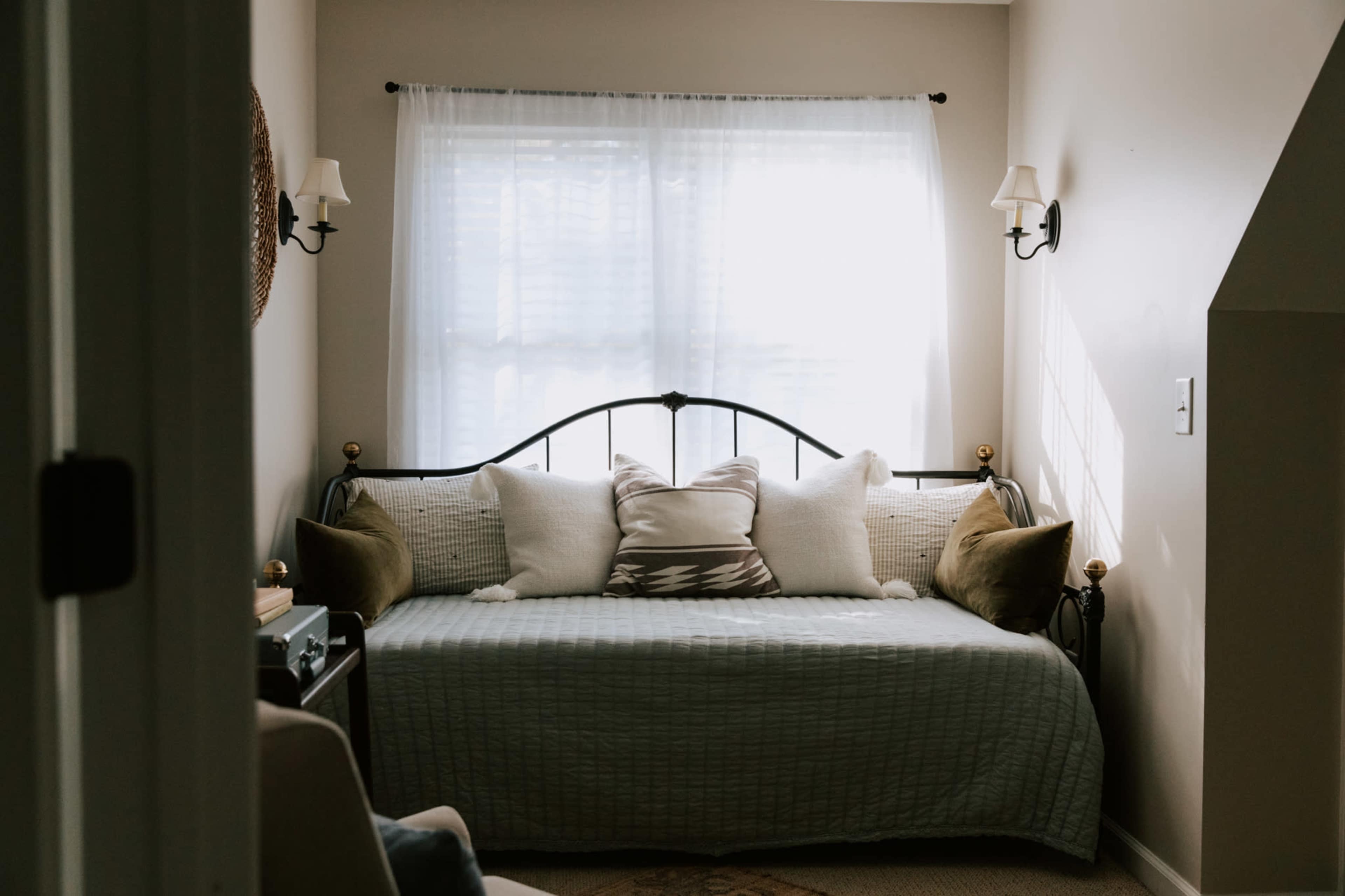 A daybed with decorative pillows is positioned against a wall under a window with sheer curtains in a softly lit room.