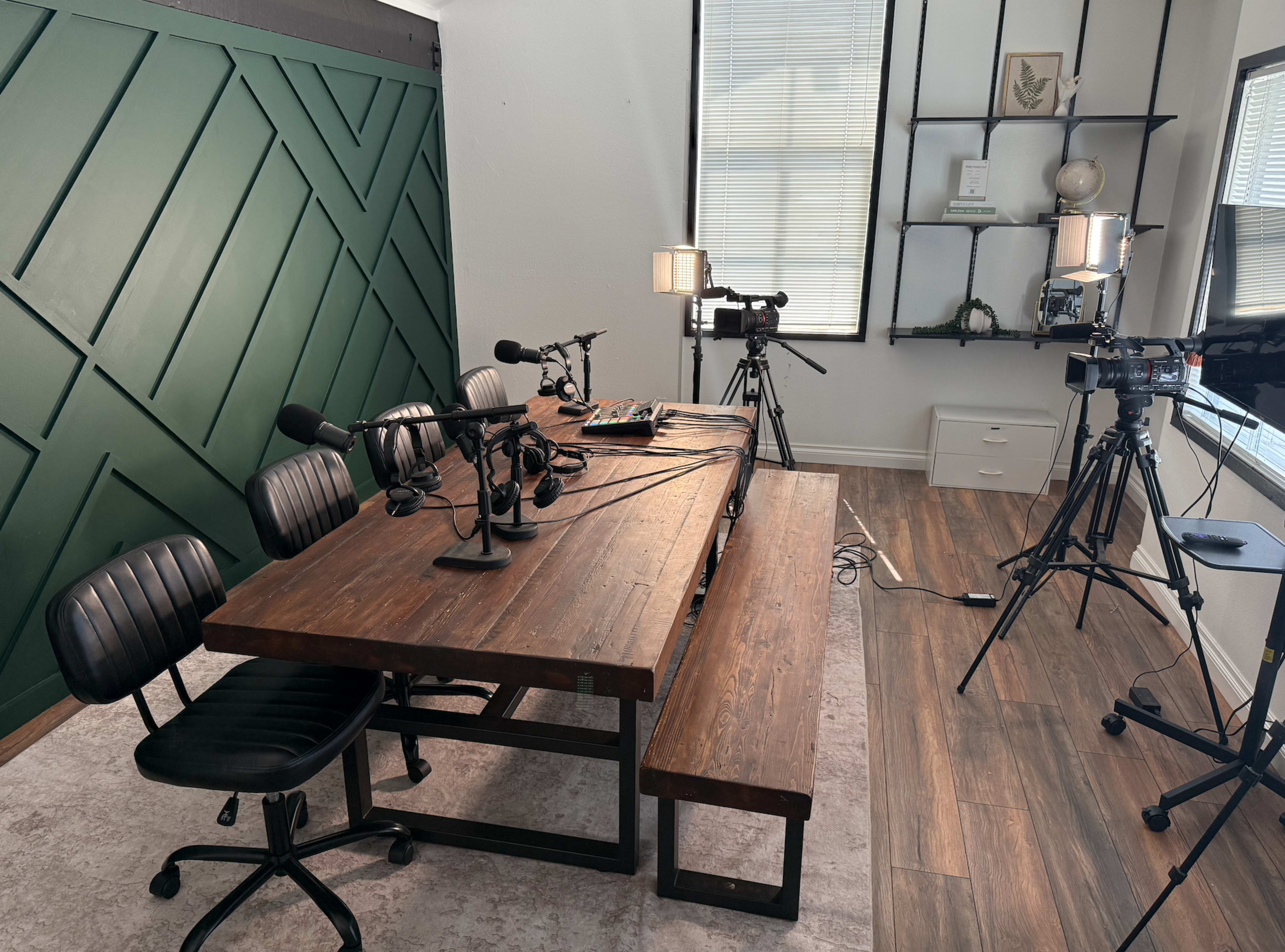 The image shows a podcast recording studio with a wooden table, several microphones, and cameras positioned around the space.