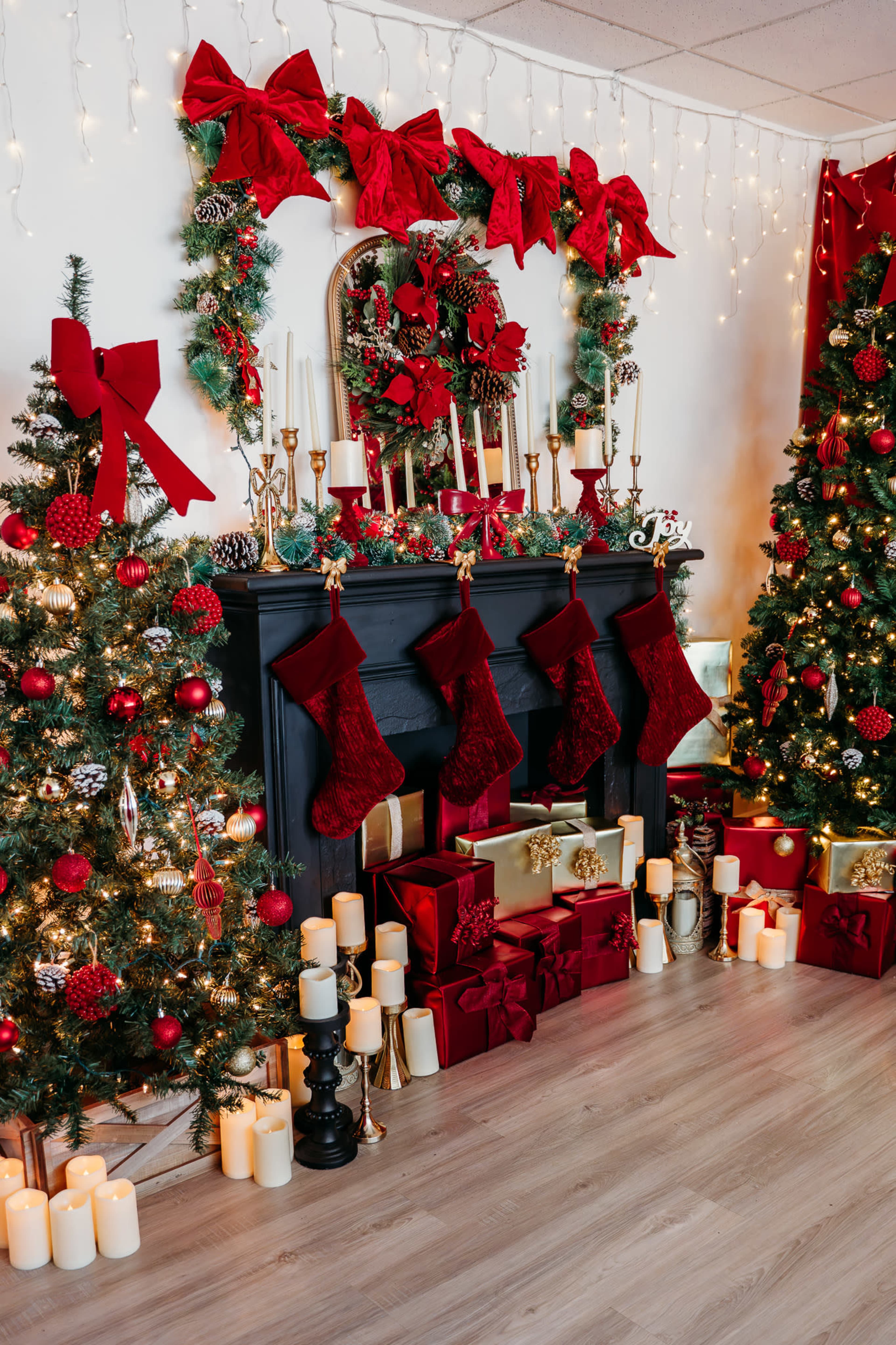 A decorated Christmas scene features a black fireplace adorned with red bows and garlands, surrounded by Christmas trees and presents, with stockings hanging above and candles lit on the floor.