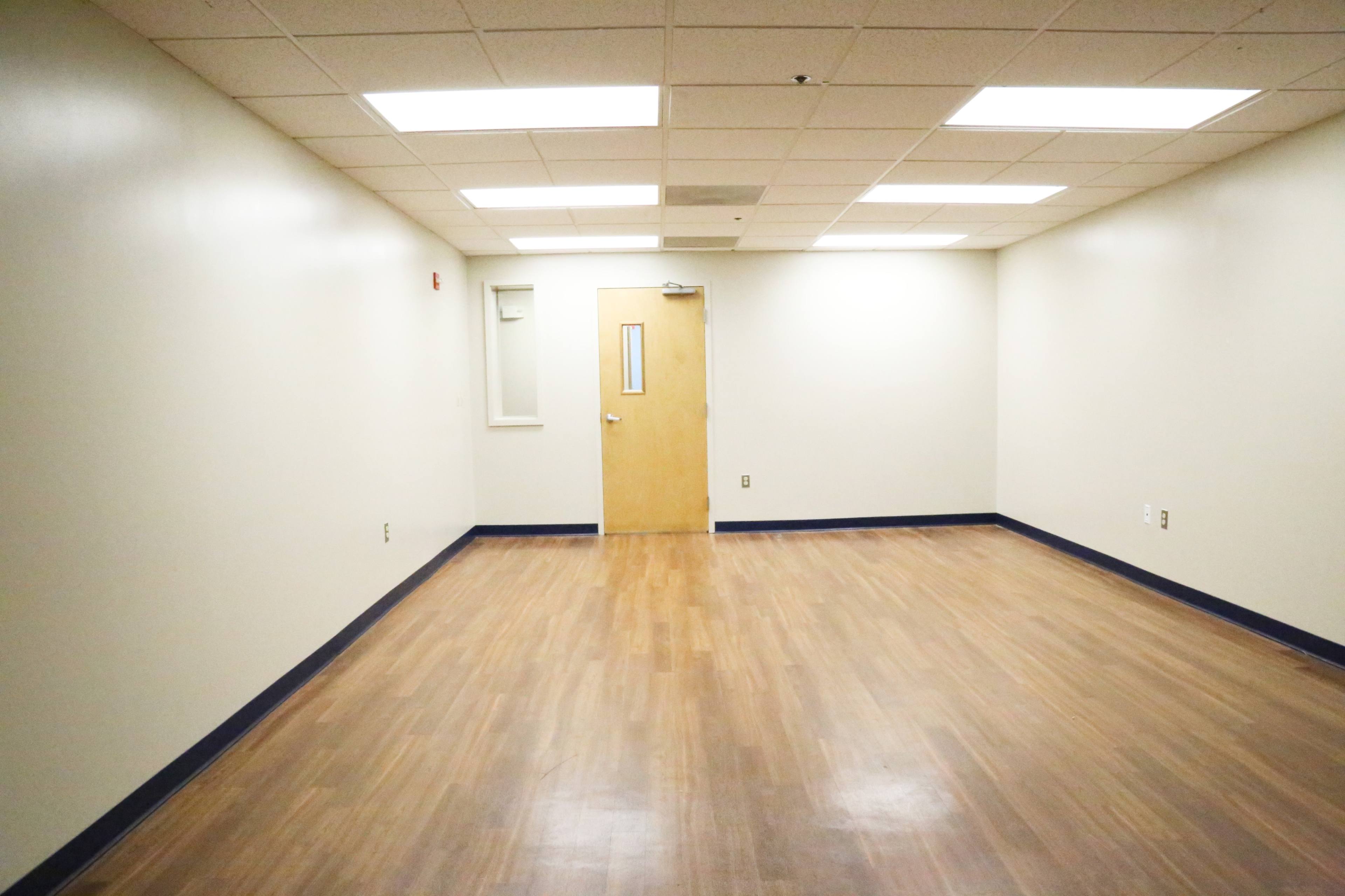 A sparsely furnished room with wooden flooring, white walls, and a single door with a small glass window.
