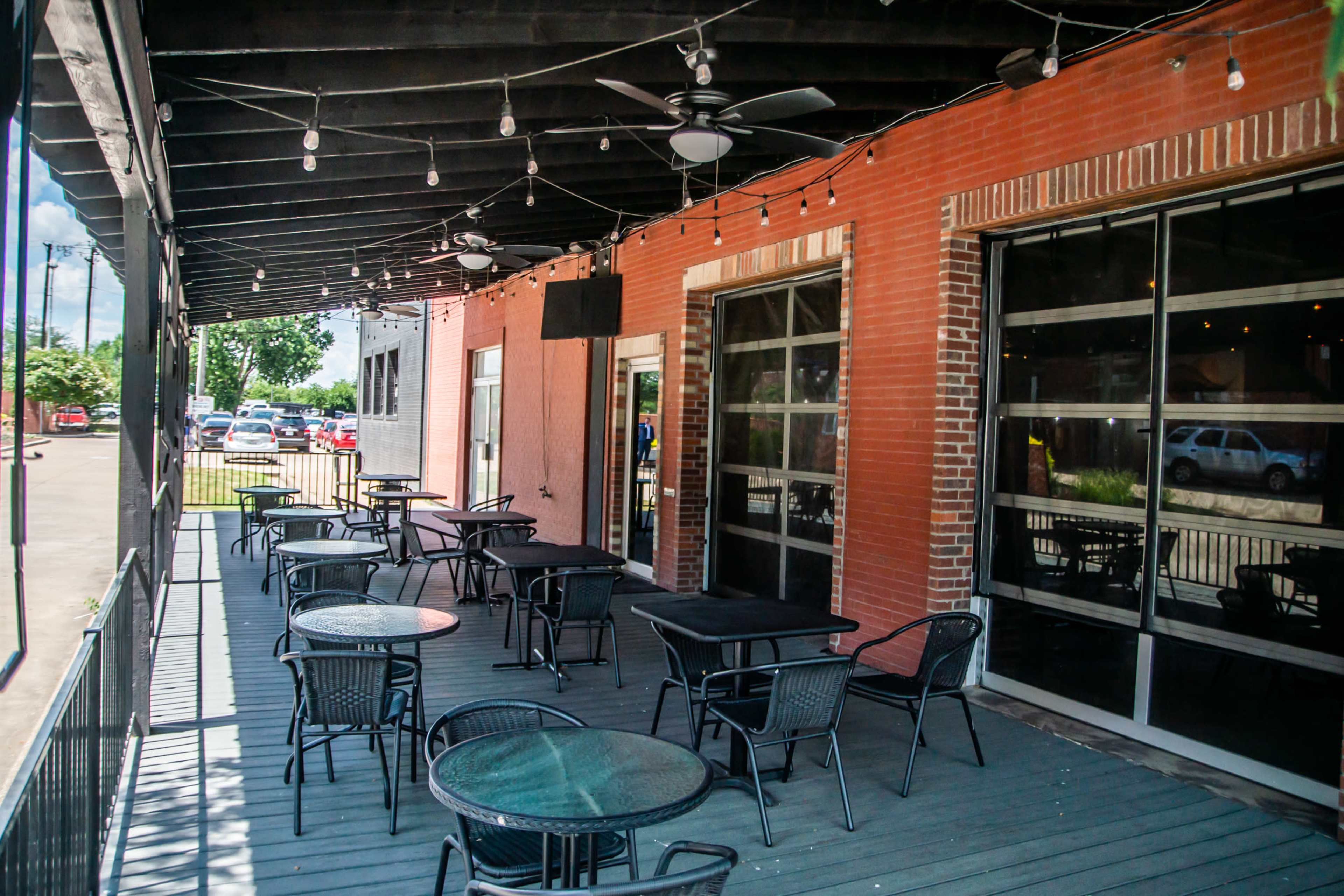 The image shows a spacious outdoor patio equipped with several black metal tables and chairs beneath a canopy with string lights.