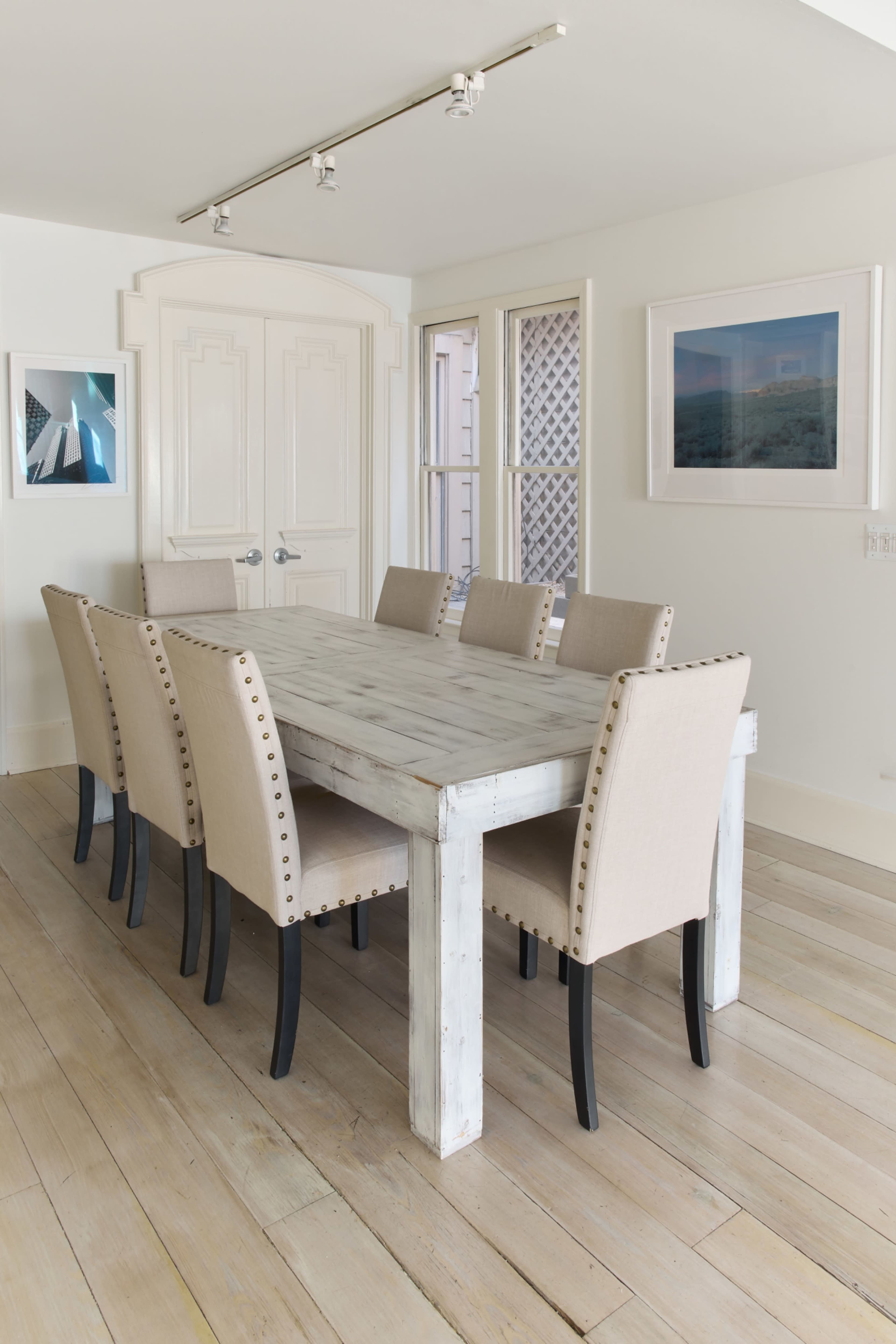 A long, wooden dining table is surrounded by eight beige chairs in a brightly lit room with white walls and large windows.