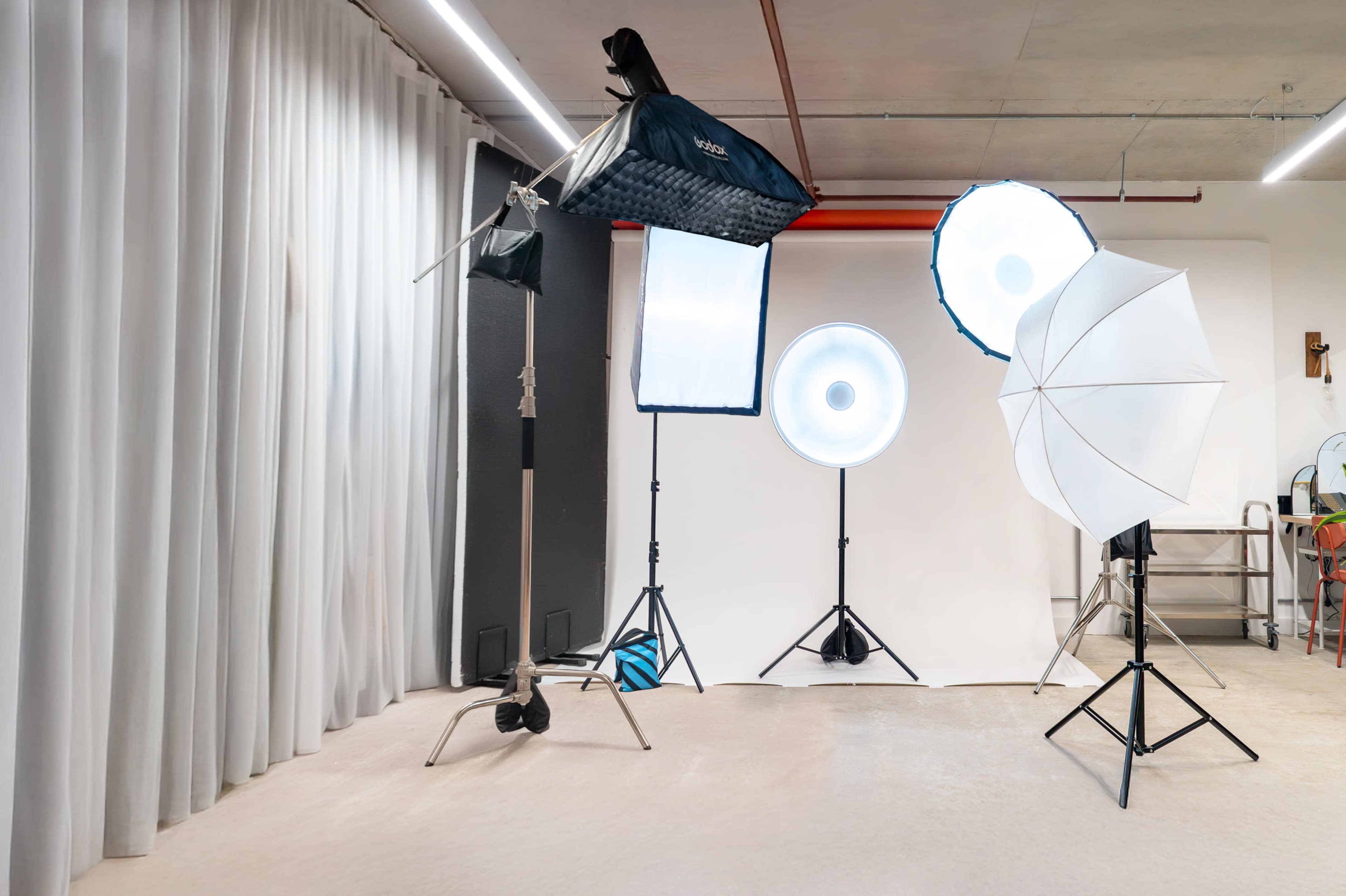 The image shows a photography studio equipped with various lighting setups, including softboxes and umbrella lights, arranged in front of a backdrop.