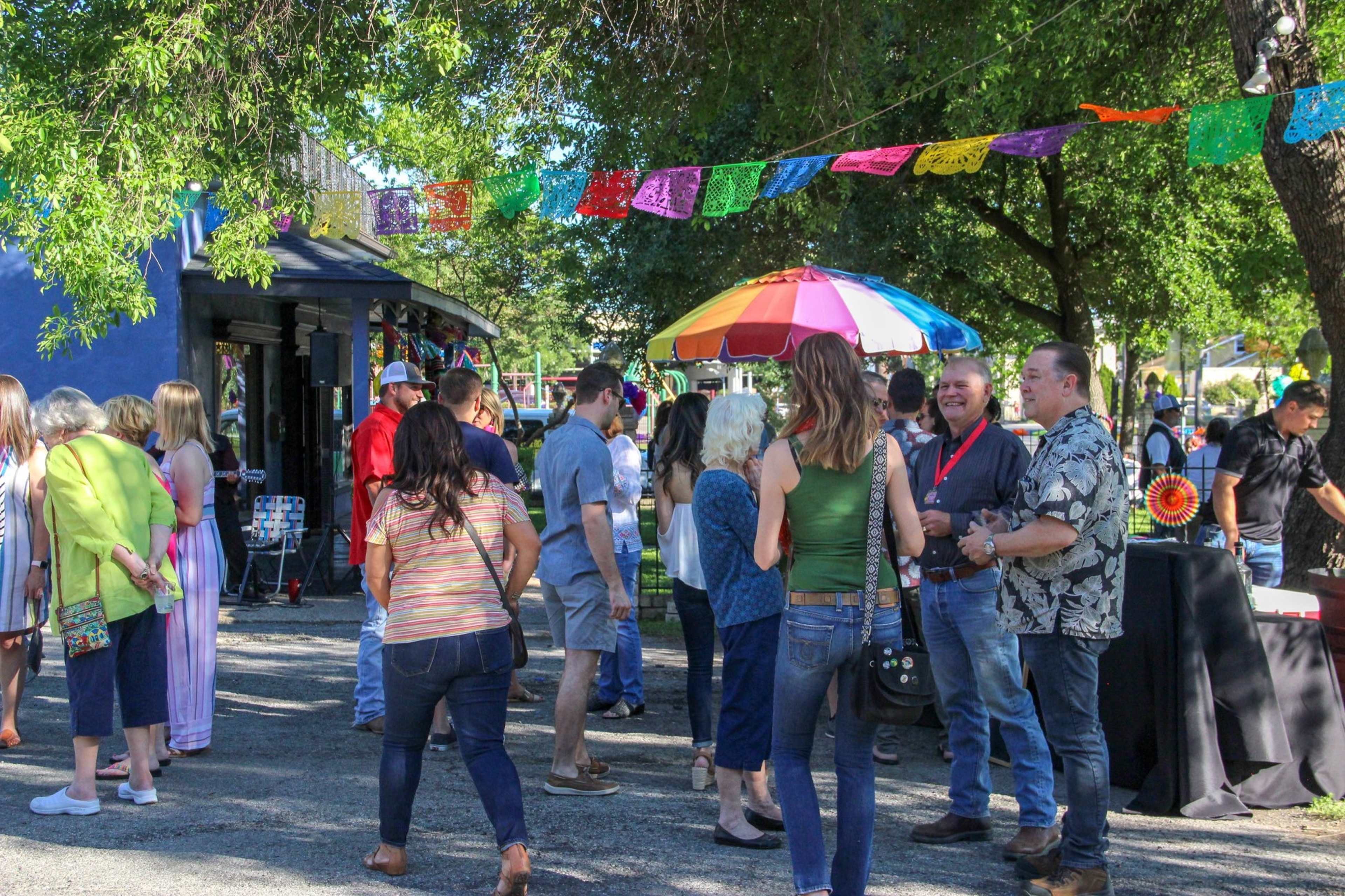 A lively outdoor event features a gathering of people conversing near colorful decorations and shaded seating.