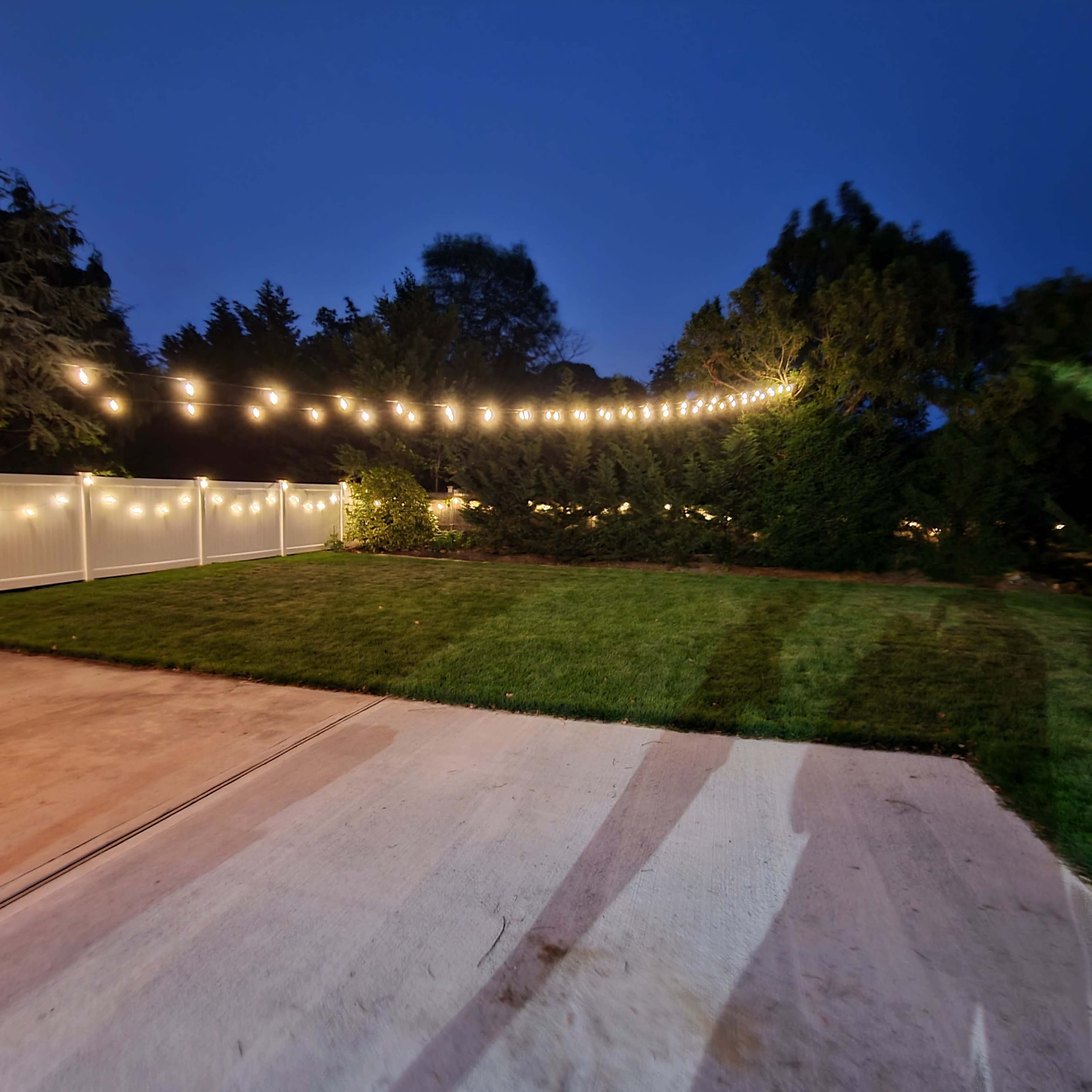 The image shows a backyard at dusk with a well-manicured lawn and string lights illuminated above a patio area.
