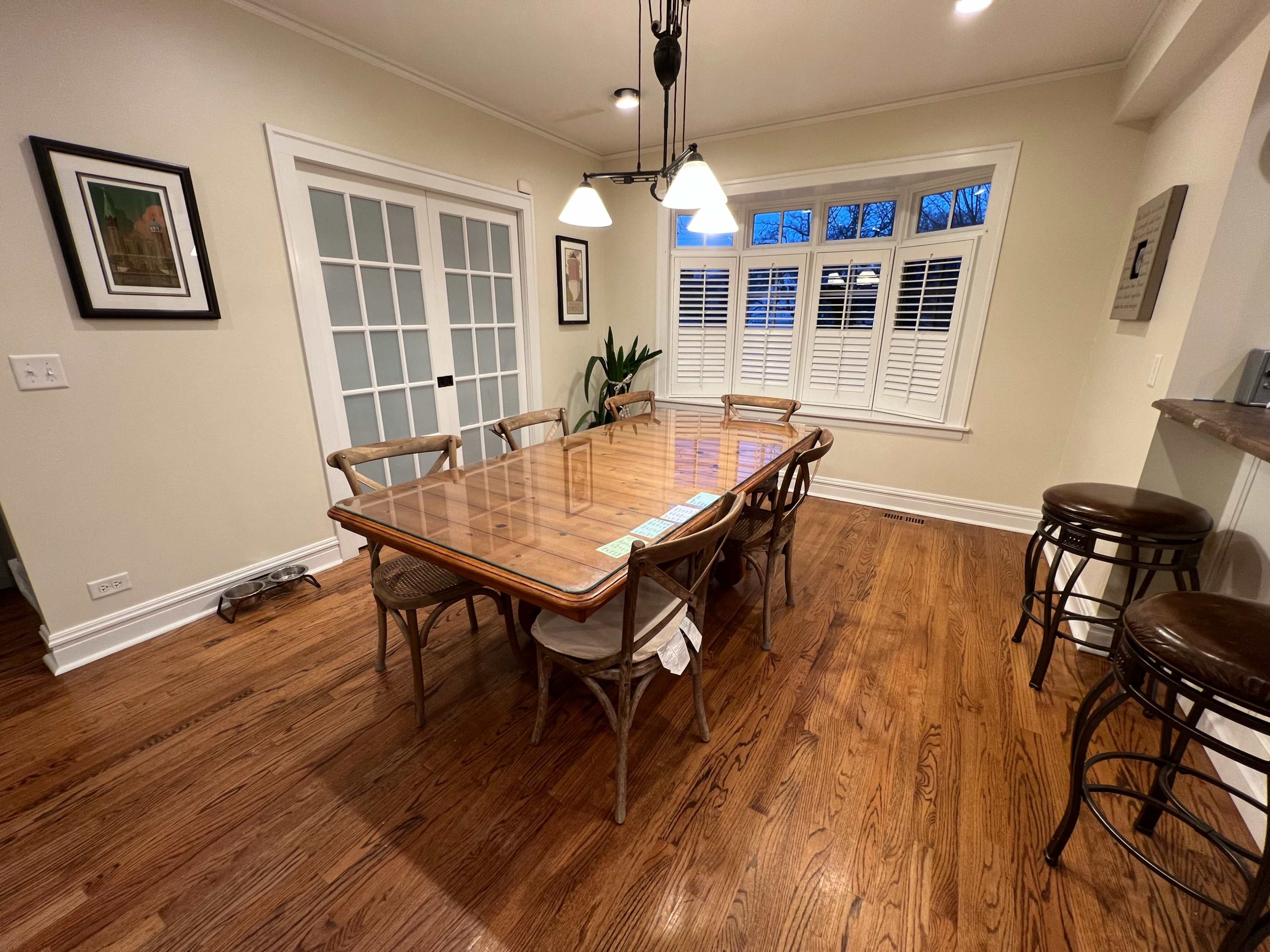 A wooden dining table is set with chairs in a well-lit room featuring large windows and a plant in the corner.